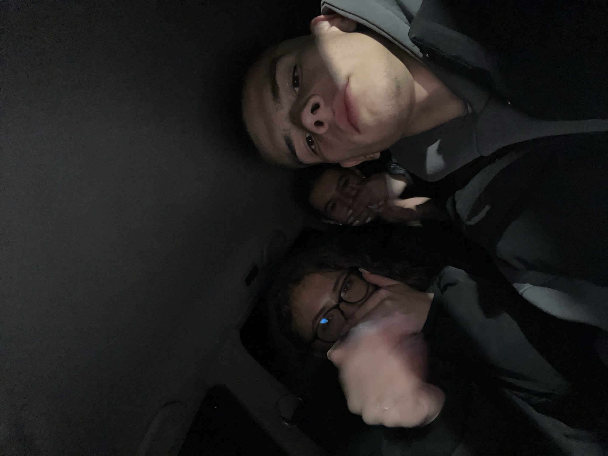 Three individuals are visible in a low-light environment, possibly the interior of a vehicle. The person in the foreground, a male with short hair, is positioned closest to the camera. Behind him, a female with glasses covers her mouth with her hand. In the background, a third person is partially obscured. The image does not provide enough information to determine its location in Karavelovo, Bulgaria.