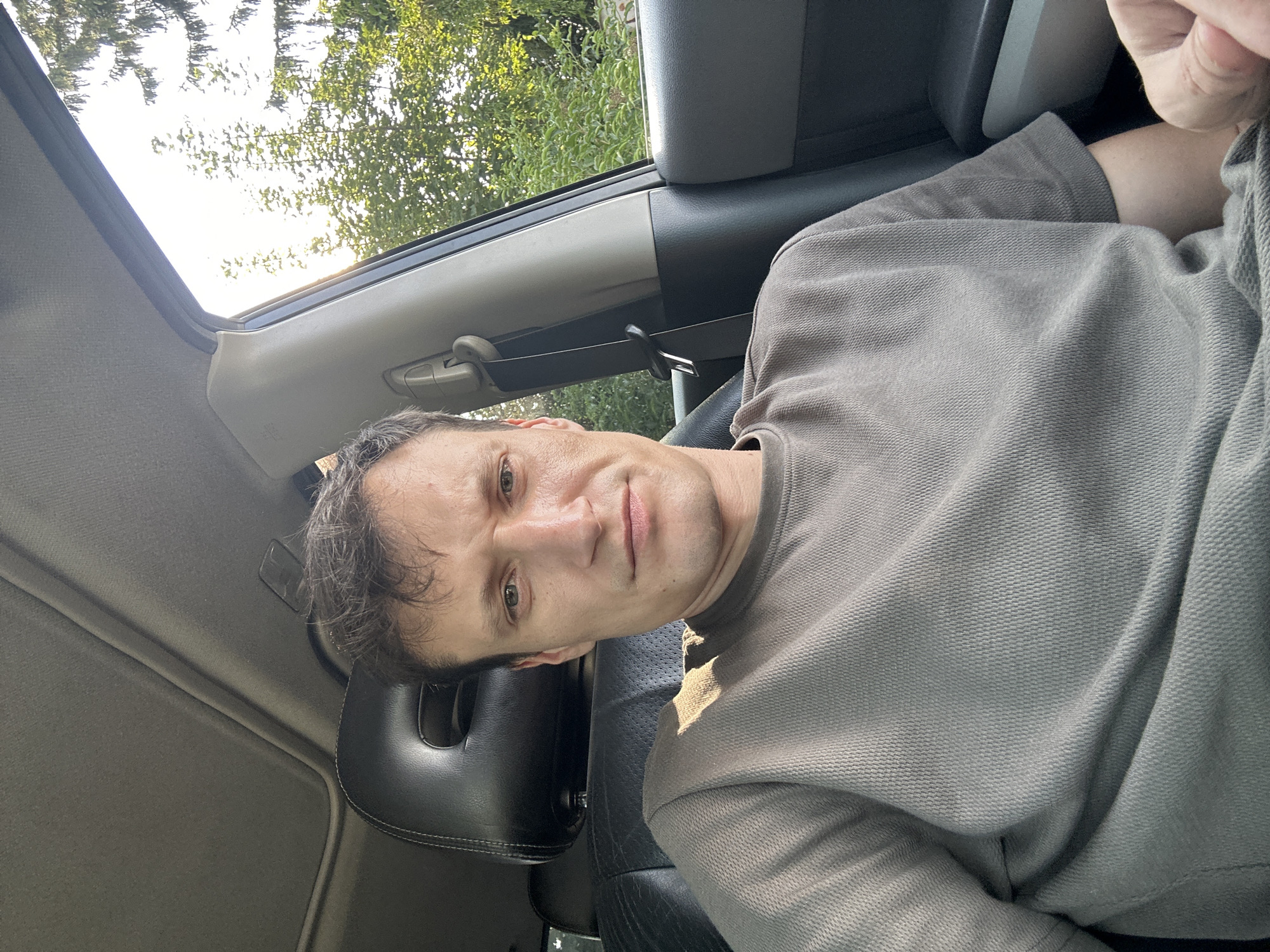 A man sits in the driver's seat of a car, looking directly at the camera. The car's interior is visible, including the black leather headrest and part of the dashboard. Sunlight streams in through the open window on the right side of the frame, illuminating the man's face and casting a shadow across his shirt. Trees and foliage are visible outside the car window, suggesting an outdoor setting. The man is wearing a gray t-shirt. The image was taken in Shatava, Ukraine.
