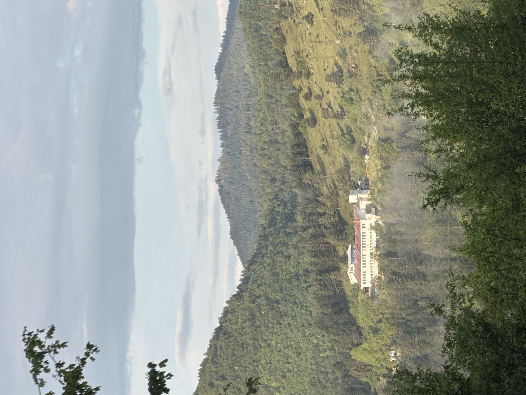 The image shows a landscape view in Strazha, Bulgaria. A white building with a red roof sits on a hillside among dense trees. The background features layers of green hills extending into the distance, with a strip of cloudy sky visible on the left. In the foreground, tree branches frame the right and bottom left of the image. The scene has a hazy, somewhat muted quality.