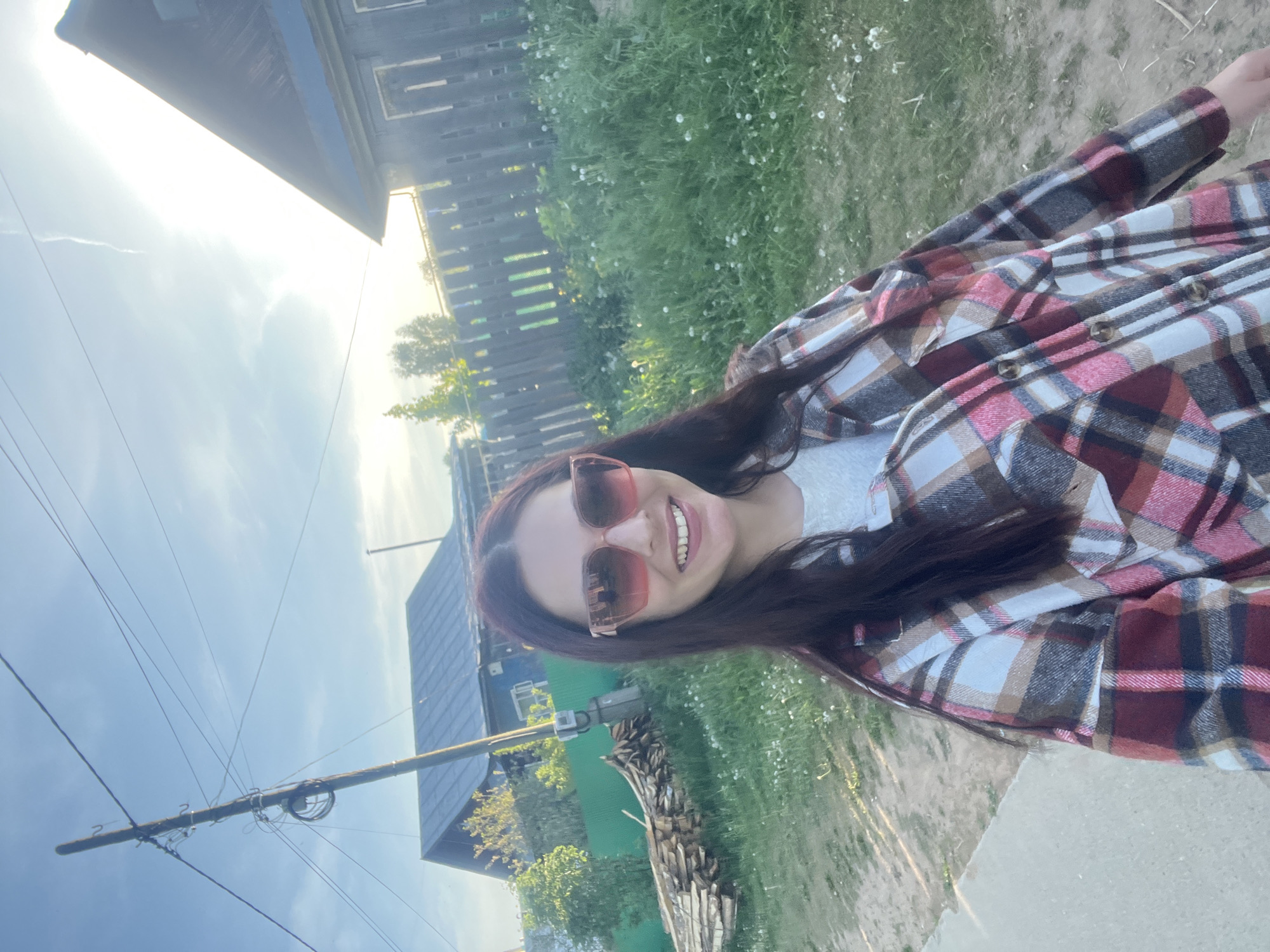 A medium shot shows a smiling woman wearing sunglasses and a plaid shirt. The image appears to be taken in Chaikovsky, Russia, based on the architectural style of the wooden buildings visible in the background. A utility pole with power lines stretches across the lower-left corner of the frame. The environment is a mix of rural and urban, with grass, trees, and flowers interspersed with buildings.