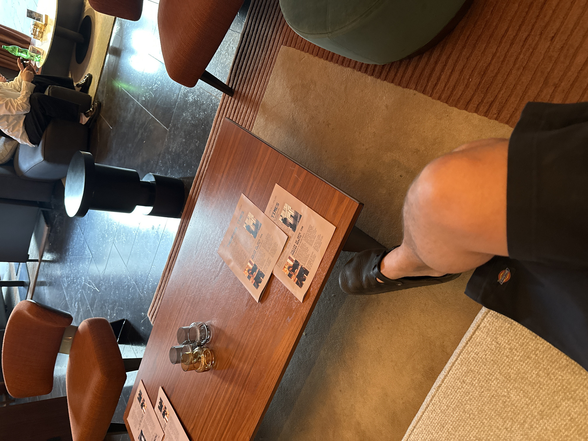 The image is a top-down shot taken indoors, likely in a café or lounge area. There is a wooden table with a few stacks of printed materials on it. One set appears to be promotional leaflets, while the other appears to be small glass containers.
A person is seated at the table, evidenced by the presence of a leg and a partially visible shoe near the edge of the shot. The person is wearing black shorts or trousers.
Furniture includes a cushioned stool in the upper right corner of the image, as well as brown chairs that are near the table.
The floor is a mix of materials, with tiling visible in the background and what appears to be a rug or carpet in the foreground.
Without more information, I cannot confirm this is taken in Madrid.