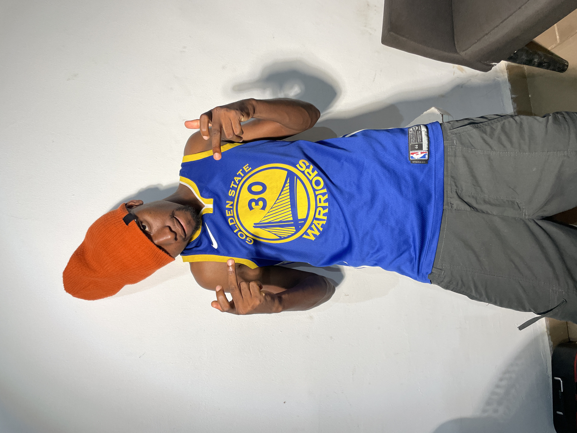 A young Black man is lying on his back against a white wall, indoors. He is wearing a blue Golden State Warriors jersey with the number 30 on it, grey pants, and an orange beanie. He is making a hand gesture with both hands. A dark gray chair is visible in the background. The location is Barakin Akawo, Nigeria.