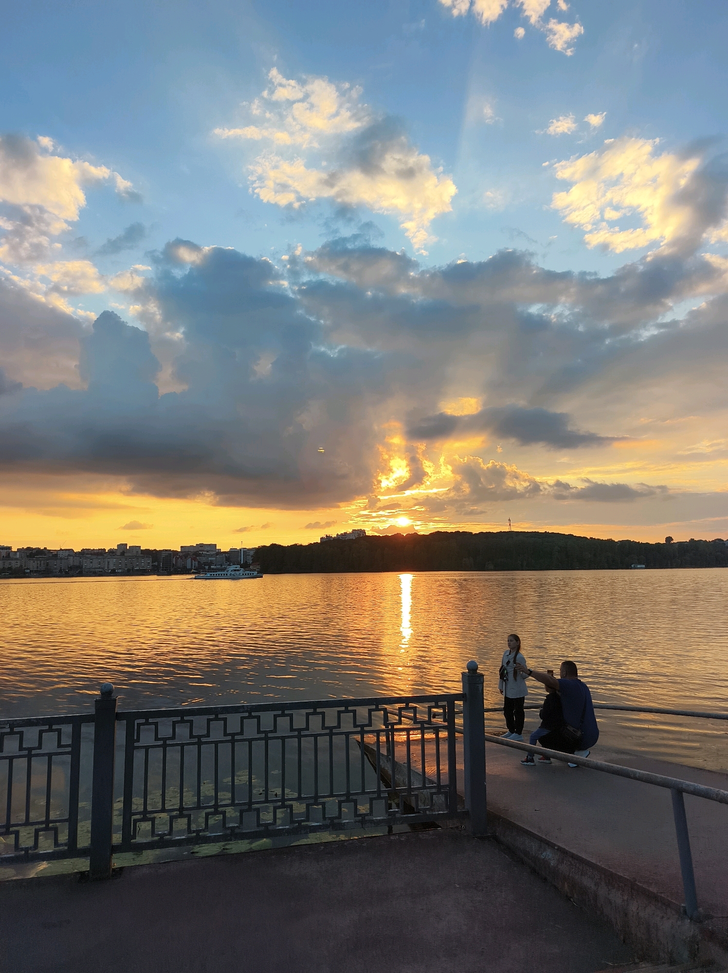 The image depicts a sunset over Ternopil Lake, Ukraine. The sky is a dramatic mix of blue and orange hues, with clouds catching the last rays of the sun. The sun itself is partially obscured by clouds, casting a golden glow across the water. A cityscape lines the distant shore, with buildings silhouetted against the vibrant sky. A white boat is visible on the water in the middle ground. In the foreground, a metal railing runs along the edge of a paved walkway that borders the lake. Two individuals, a woman and a child, are standing near the railing, with a man crouching beside them. They appear to be enjoying the view and possibly taking photographs. The water is calm, with gentle ripples reflecting the warm colors of the sunset. The overall impression is one of tranquility and natural beauty at the close of day.