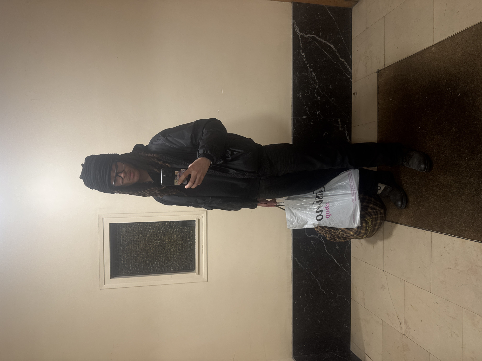 A single individual stands centered in an indoor hallway, consistent with a residential building in Paris, France. The person wears a black head covering, dark-rimmed glasses, a black bomber jacket, black trousers, and dark shoes; dreadlocks are visible. They hold a dark smartphone, adorned with multiple stickers, at chest level, appearing to operate it. In their left hand, they carry a translucent white plastic shopping bag displaying the reversed "Casino" logo, positioned above a brown and tan checkered bag. The background features light beige walls with a dark marble wainscoting. A small, framed window with textured glass is present on the left wall. The floor consists of light-colored rectangular tiles, with a dark brown doormat in the immediate foreground.