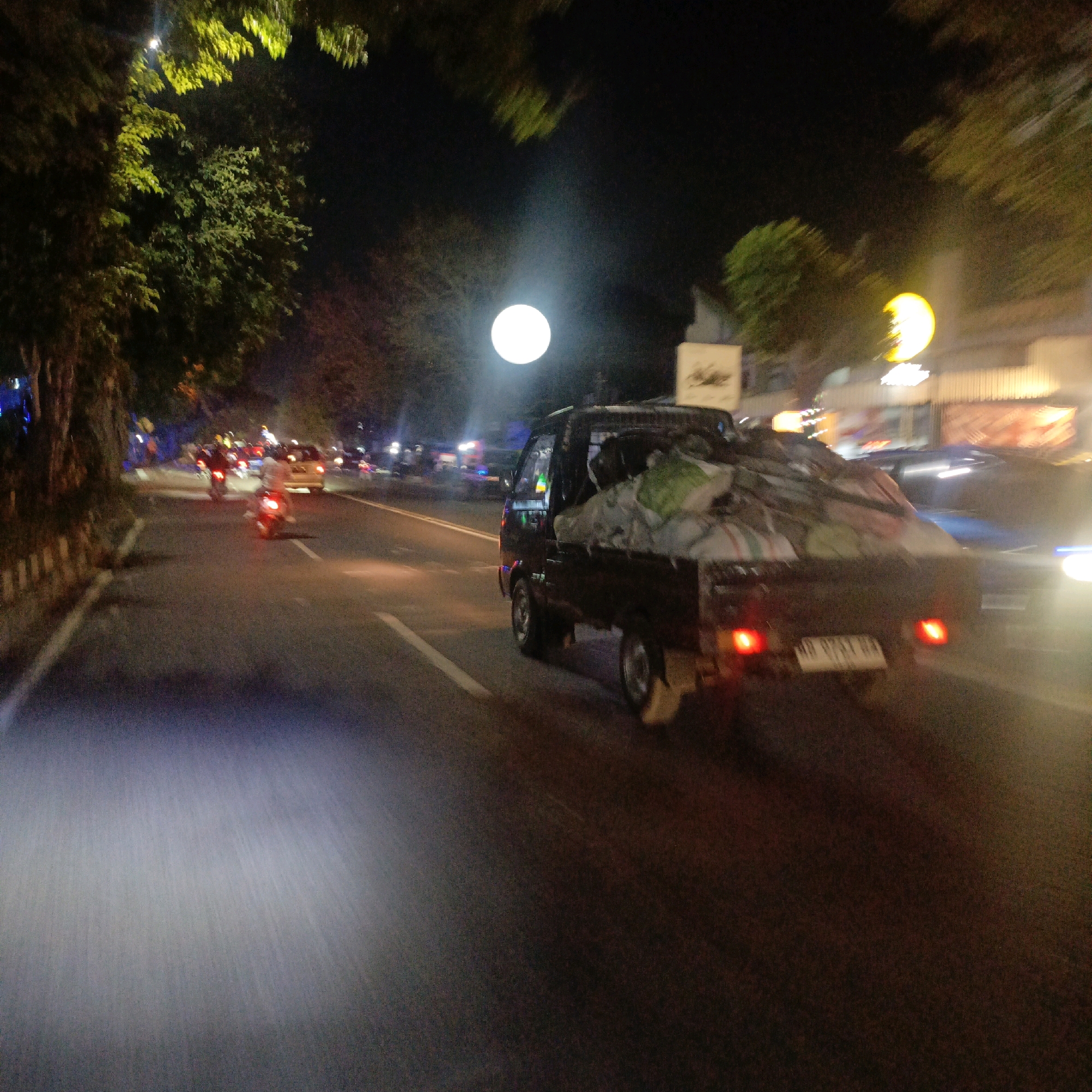 An open-bed pickup truck, laden with what appears to be large bags of produce covered in tarpaulin, travels down a street at night in Boyolali, Indonesia. The truck's brake lights are illuminated. The street is asphalt with white lane markings. The scene is illuminated by streetlights and the headlights of passing vehicles, creating a blurred effect due to motion. Trees line the left side of the road, their leaves catching the light. In the background, other vehicles, including motorcycles and cars, are visible, as are illuminated signs and buildings. The overall atmosphere is that of a busy, well-lit urban thoroughfare at night.