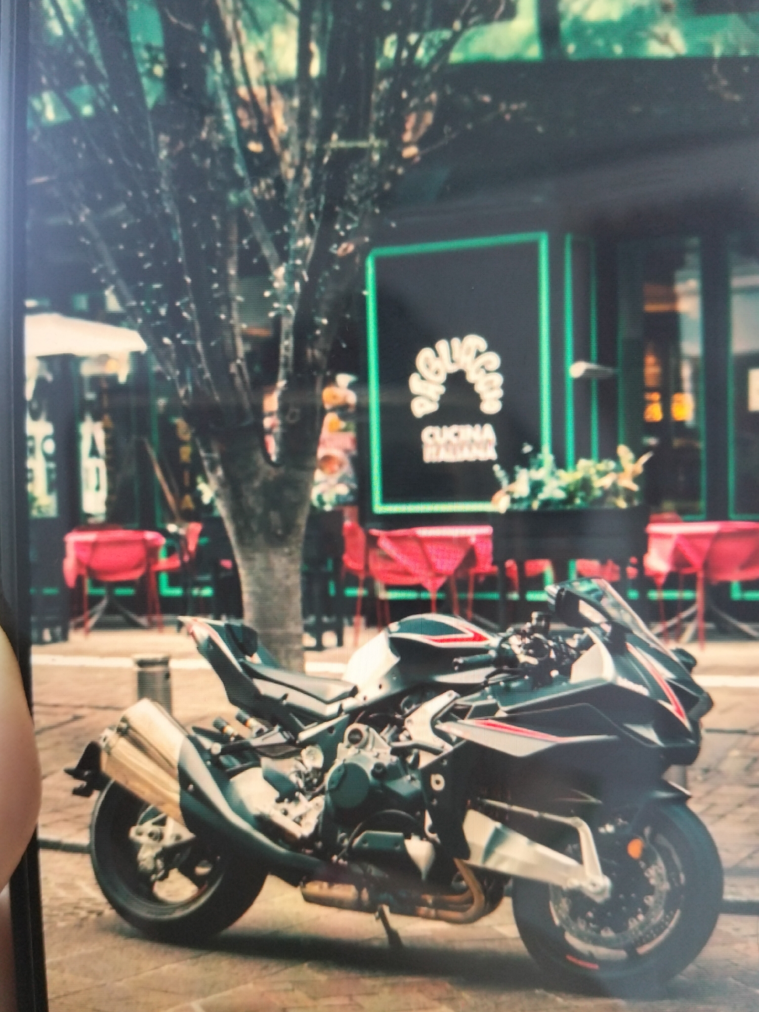 The image depicts a street scene in what is identified as Payagaboh, Indonesia. In the foreground, a black and white motorcycle is parked. The motorcycle has red accents on its body. Behind the motorcycle, a restaurant with a dark facade is visible. The restaurant features a sign with green neon outlines that reads "RAGIO" in an arched font above "CUCINA ITALIANA". There are red tables and chairs set up outside the restaurant. A large tree with dark branches dominates the middle ground. The background shows more of the restaurant's interior and other establishments. The overall lighting suggests it might be daytime, possibly overcast. No people are clearly discernible in the image.