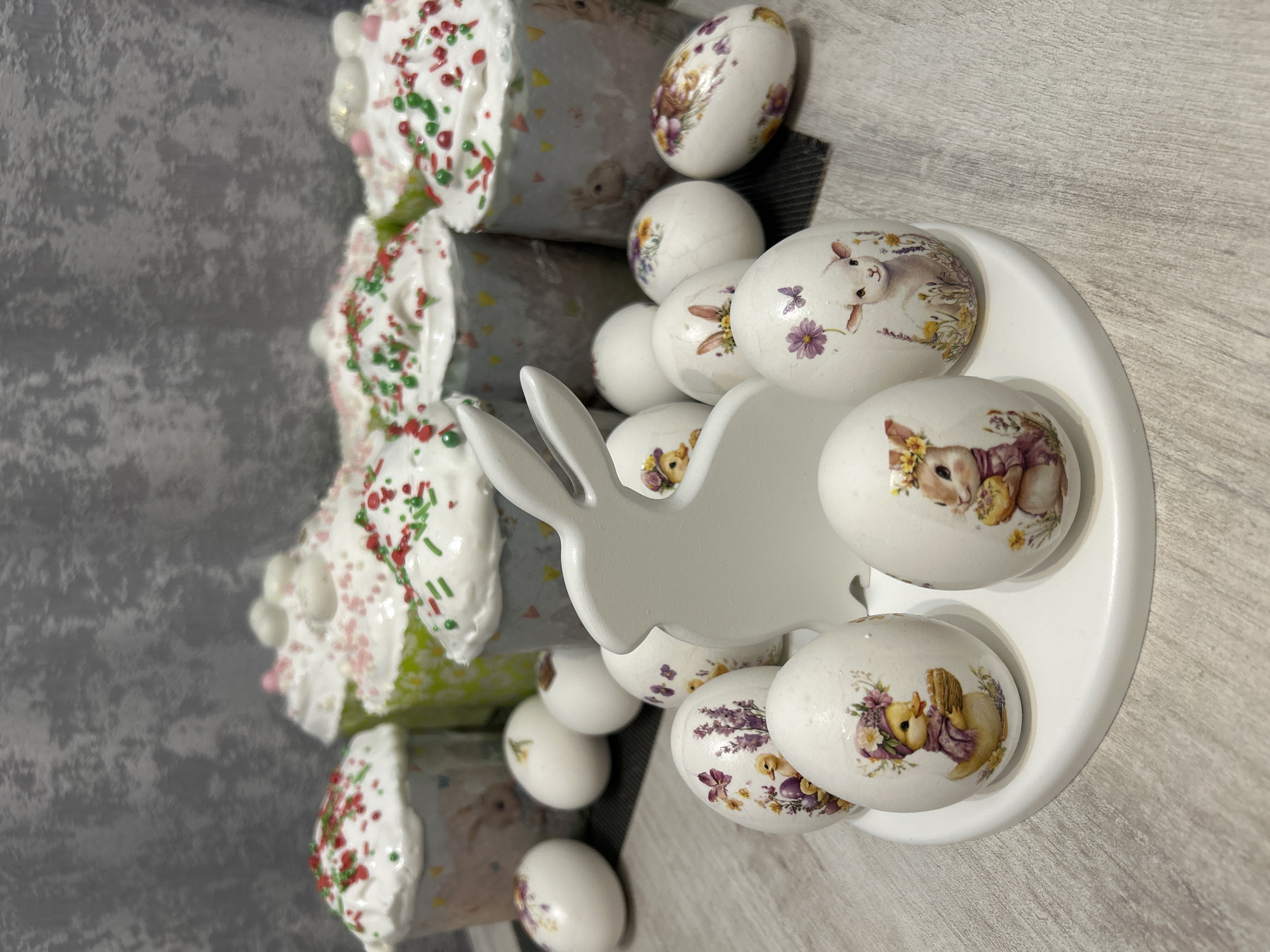 The image features a tabletop Easter display with four Easter cakes (kulich) topped with white icing and colored sprinkles, positioned on the left side of the frame. To the right, there's a white plate holding decorated Easter eggs, most of which feature images of rabbits, flowers, and chicks, applied via decoupage. A white rabbit figurine stands among the eggs on the plate. The background is a mix of gray and light gray surfaces.
The image does not provide enough information to confirm the location as Klin, Russia. This would require further context or specific details within the image linking it to Klin.