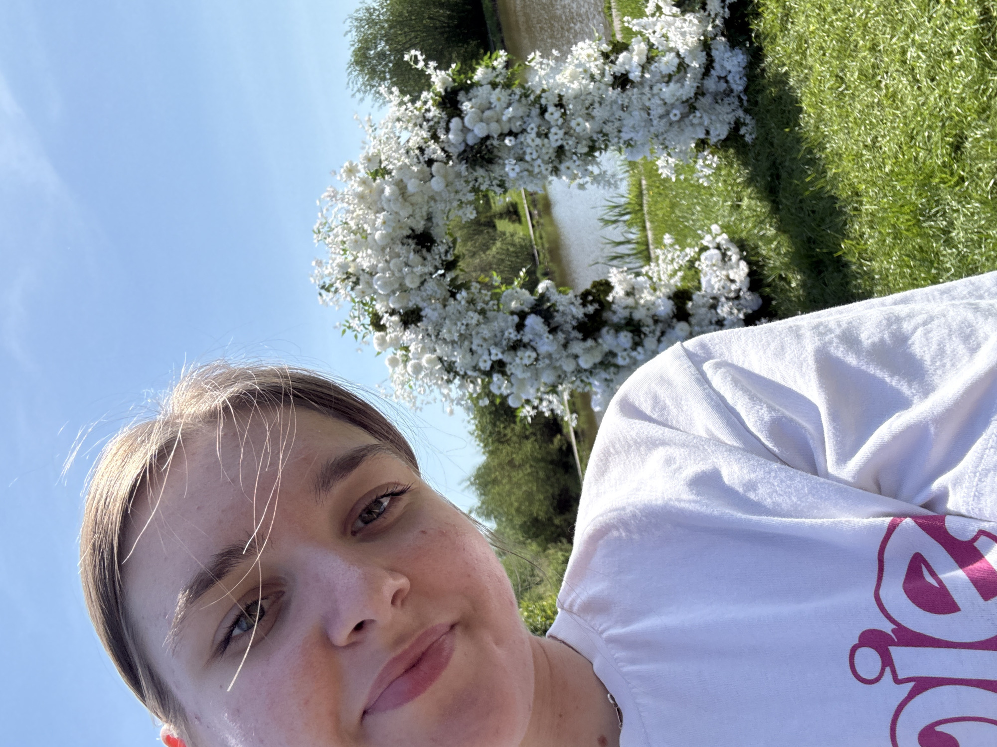 A selfie image shows a person with fair skin, light brown hair, and blue eyes. The subject is wearing a white t-shirt with a pink design. The person appears to be outdoors on a sunny day. In the background, there is a circular floral arrangement of white flowers, possibly for a wedding ceremony. Beyond the flowers, there is a body of water and green grass. Based on the image's geolocation metadata, the location is in Нижние Муллы, Russia.