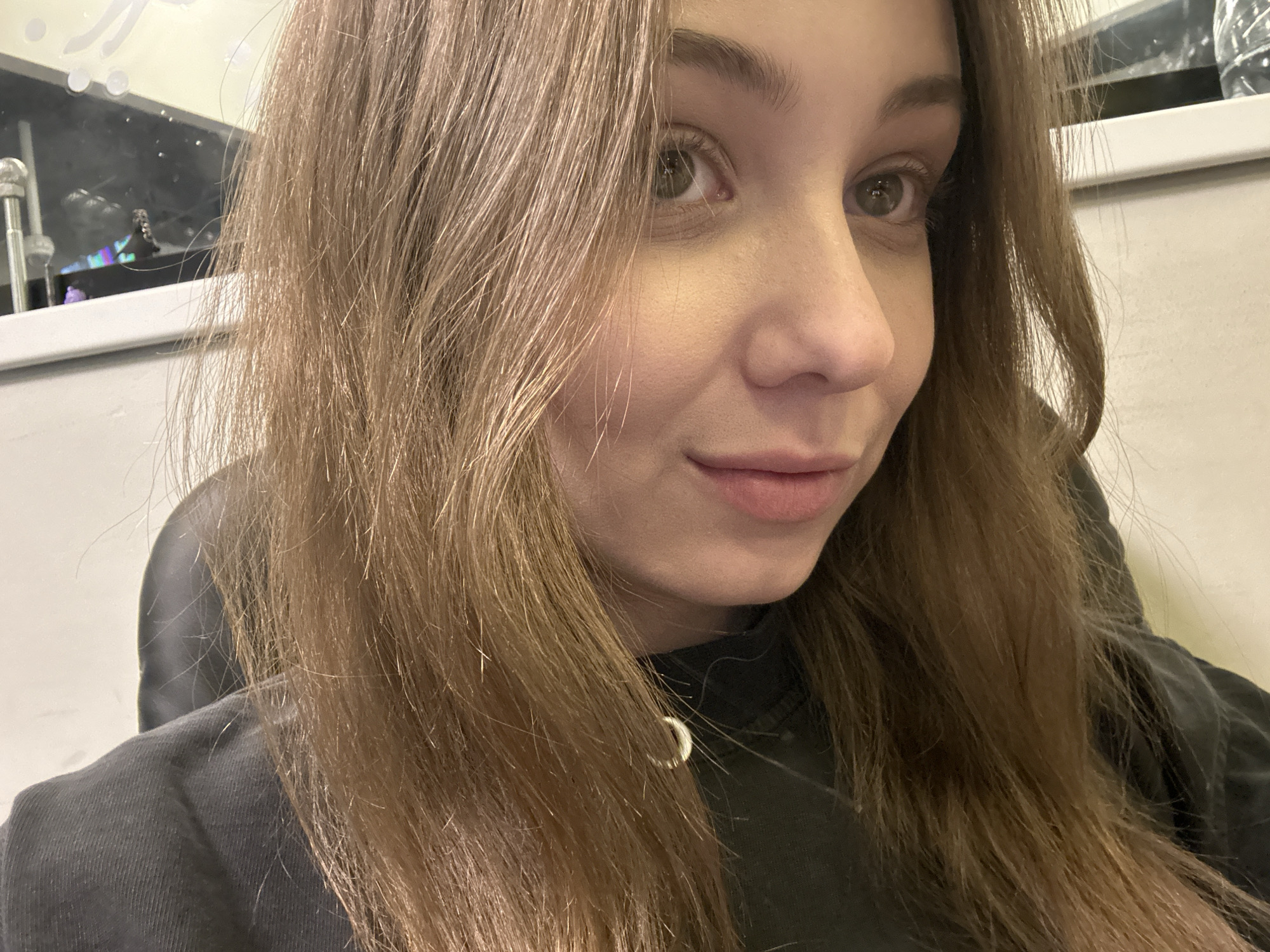 A close-up selfie shows a young woman with light brown hair and fair skin. She has light green eyes and a slight smile. She is wearing a dark grey top with a silver pendant necklace. The background includes a window ledge with miscellaneous items. The location is not discernible as being in Khimki, Russia based on the provided image.
