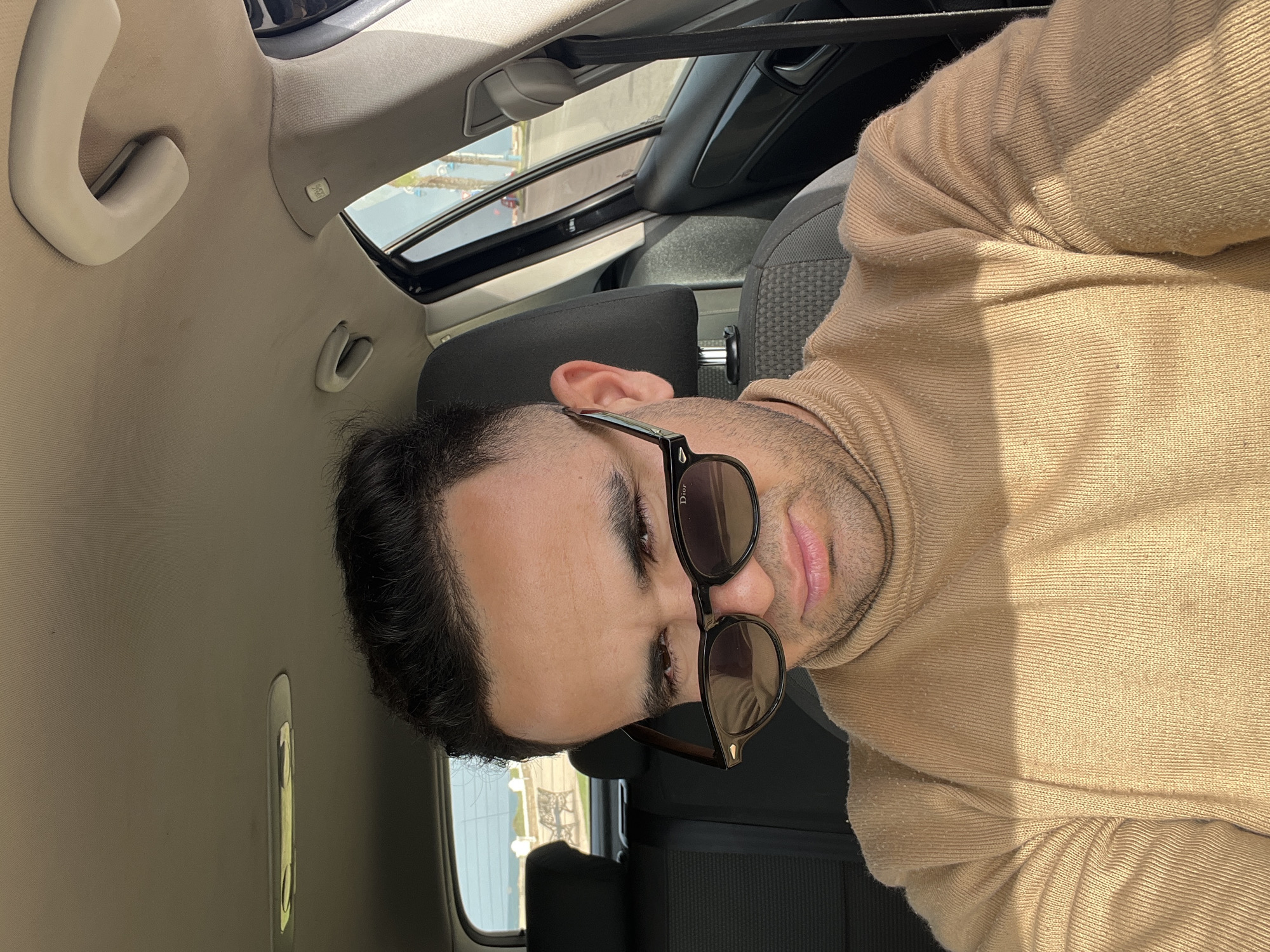 A medium close-up shot, rotated 90 degrees clockwise, features a man inside a car on a sunny day. The man, with dark hair and facial stubble, wears dark sunglasses and a light beige turtleneck sweater. The brand name "Dior" is visible in white lettering on the right arm of his sunglasses. He gazes slightly to his left, with a neutral expression.
The setting is the interior of a car, illuminated by bright natural light streaming in, indicating daytime. A sunroof is partially visible in the upper right background, contributing to the bright lighting and casting some shadows. The car's headliner is a light-colored fabric, and a beige grab handle is prominent in the upper left. The seats appear to be dark grey. Through the car window in the lower left, a glimpse of the outside world shows what appears to be a large body of water and a distant metal structure, suggestive of a coastal location. The man's knitted sweater exhibits slight pilling on the fabric. The image conveys a casual and relaxed moment.