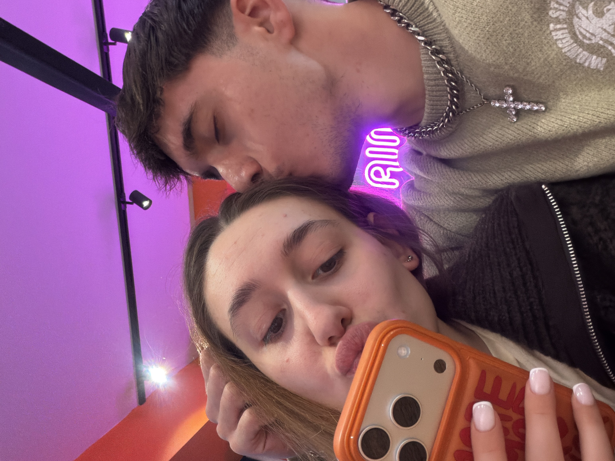 A young man kisses a young woman's forehead while she takes a selfie with an orange phone case. The man wears a silver chain with a cross pendant and a light-colored sweater. The woman has manicured nails with white tips. The background includes a black metal structure, track lighting, and a purple neon sign that reads "RIO". Based on the presence of the "RIO" neon sign and the interior design, the location is likely the Rio Rooftop Bar in Sofia, Bulgaria.