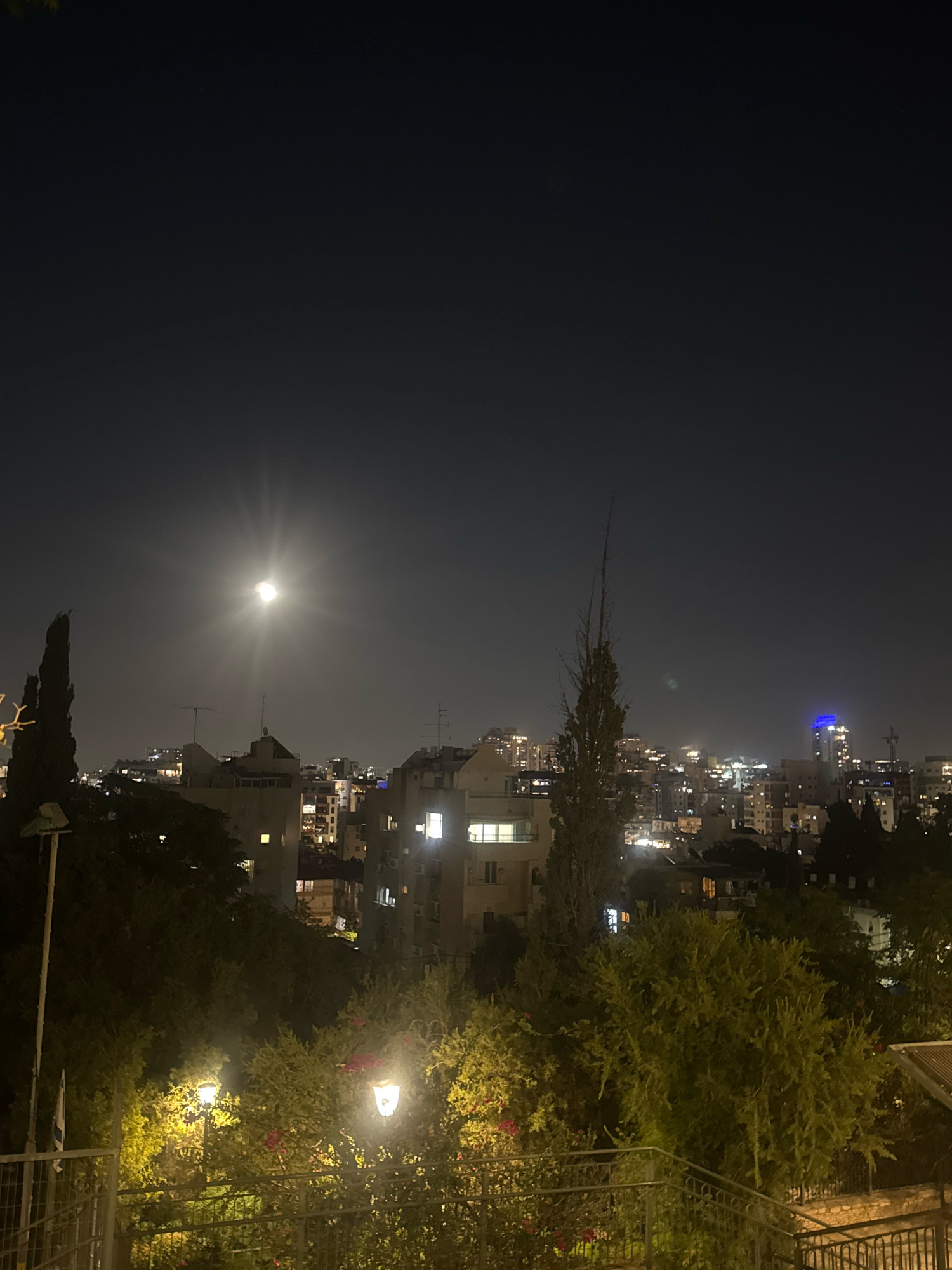 A nighttime cityscape of Ramat Gan, Israel, is depicted. The sky is dark with a bright full moon positioned towards the left of the frame, casting a distinct halo. The moon illuminates a dense urban landscape filled with buildings of varying heights, many of which have illuminated windows. In the foreground, lush green trees and bushes are visible, with two streetlights casting a warm yellow glow on the foliage. A metal railing runs along the bottom of the frame, suggesting a viewpoint from a balcony or elevated area. The overall scene conveys a peaceful, moonlit night in a bustling city.
