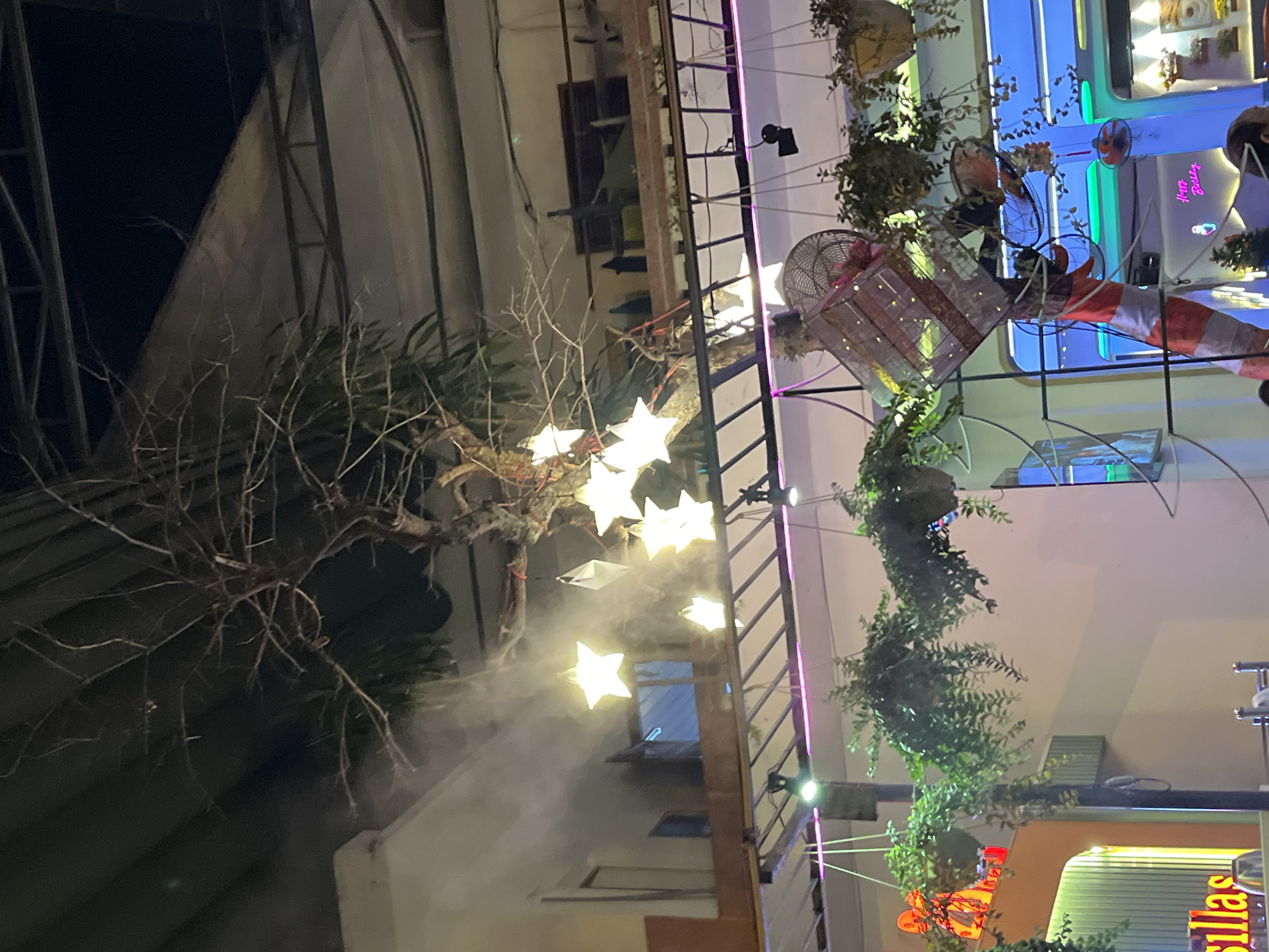 The image depicts an outdoor scene at night, likely a restaurant or cafe, in Nha Trang, Vietnam. A bare tree with intertwined branches is adorned with several illuminated star-shaped lanterns and some white paper decorations. A fine mist is being sprayed, creating a hazy effect around the lanterns and parts of the tree. Below the tree, there is a multi-level structure with hanging plants in pots. On the lower level, a decorative element resembling an oversized, upturned fan cage is visible. Further in the background, neon lighting from signs is apparent, with one sign reading "Happy Birthday." The overall ambiance suggests an evening gathering or dining area.