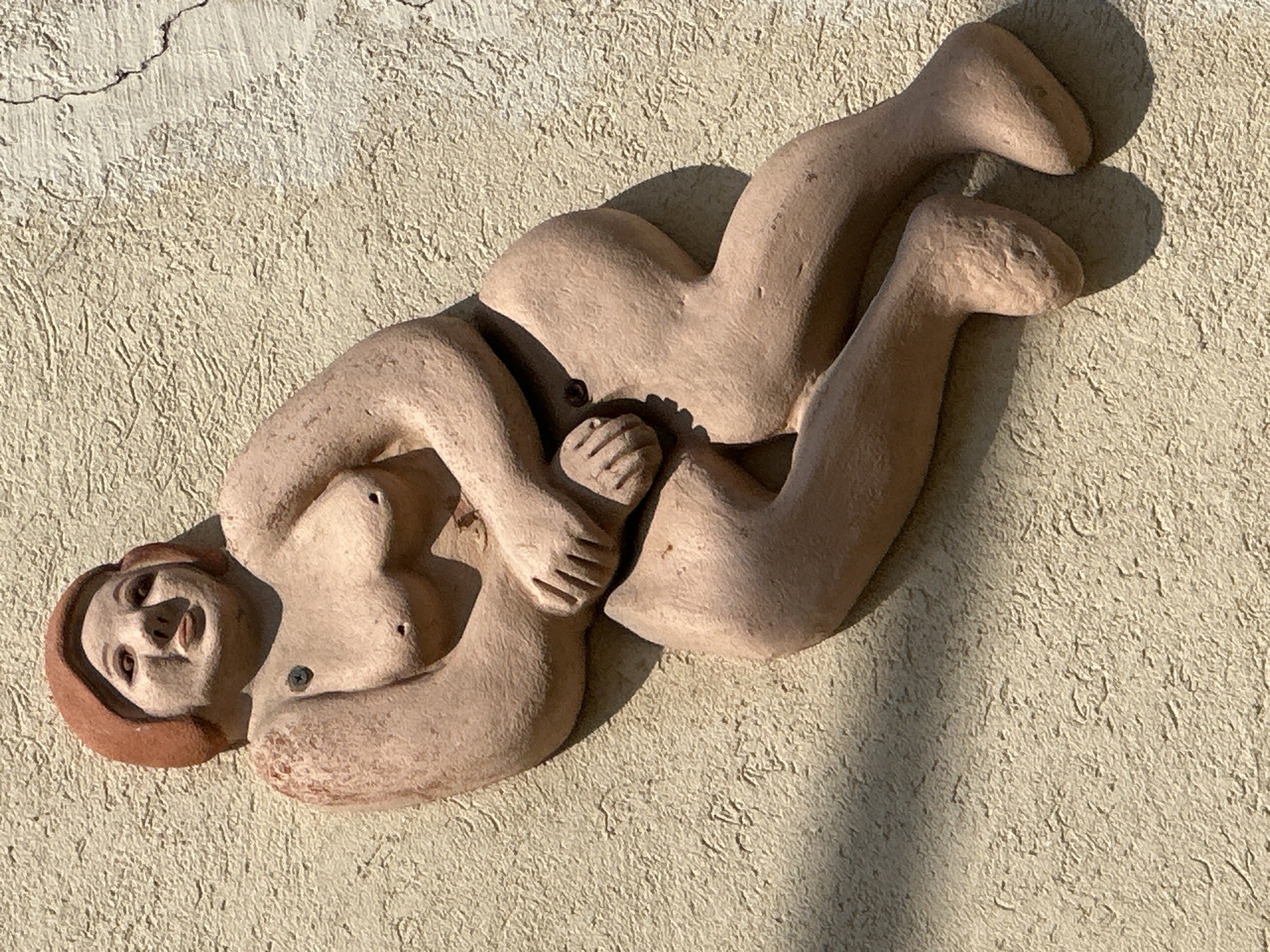 The image shows a close-up, high-angle, sunlit photograph of a terra cotta relief sculpture of a nude woman, attached to a textured, light-colored stucco wall. The sculpture depicts the woman reclining, with her legs bent and crossed, and her arms clasped in front of her. Her head is turned to the left, with her face looking slightly upwards and to the left. The wall has some visible cracks. A shadow from an unseen object falls across the lower left portion of the sculpture and the wall. The sculpture is attached to the wall with what appears to be screws. The style of the sculpture is somewhat stylized and rounded. This artwork is located on the exterior wall of a building at 40 Ben Yehuda Street in Tel-Aviv, Israel.