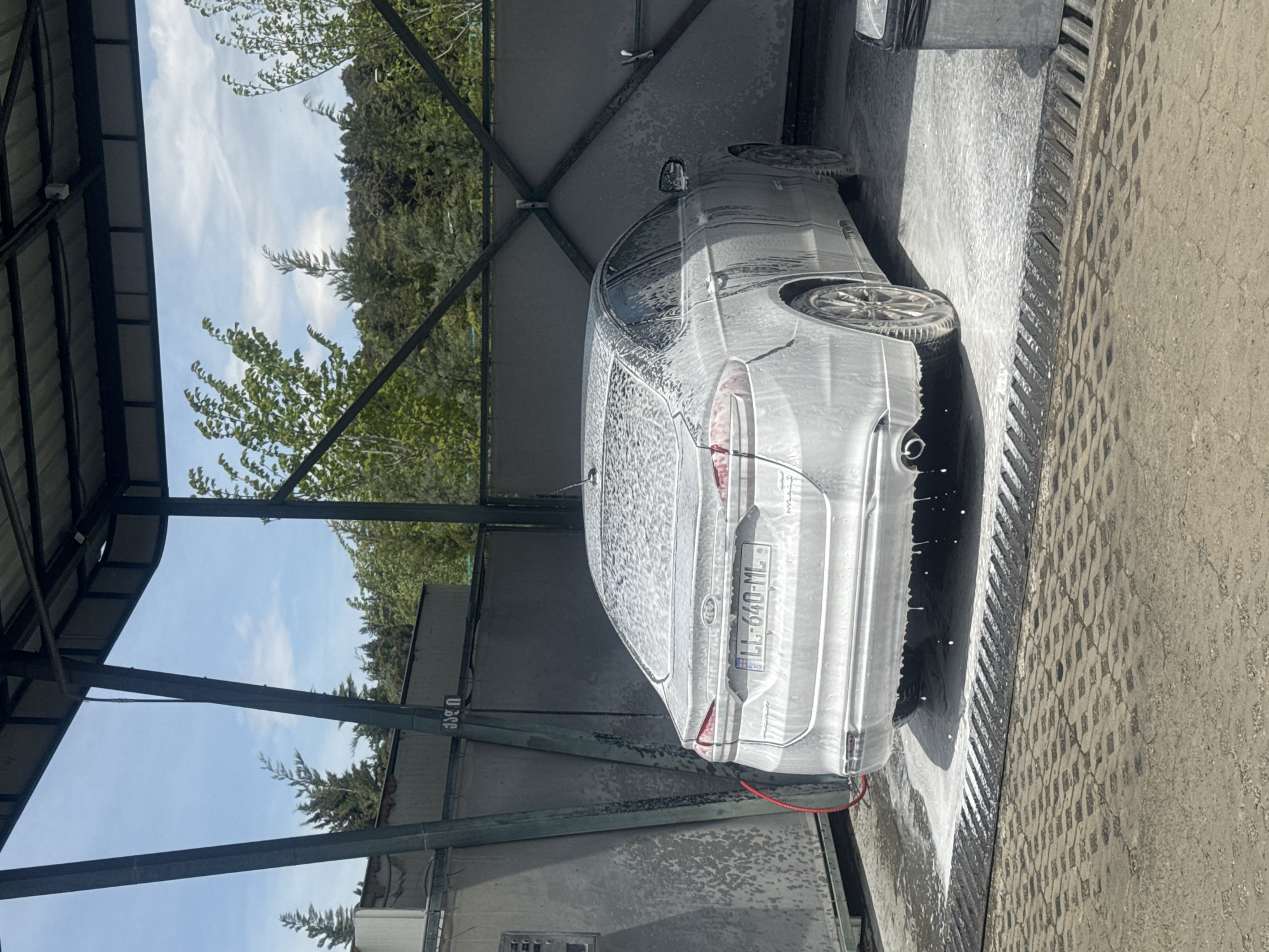 A car is being washed in a car wash, probably in Tbilisi, Georgia. The car is silver and covered in soap. It is parked next to a drain. The car's license plate is LL-640-ML. The car wash has a metal frame. The background shows trees and a blue sky with some clouds.
