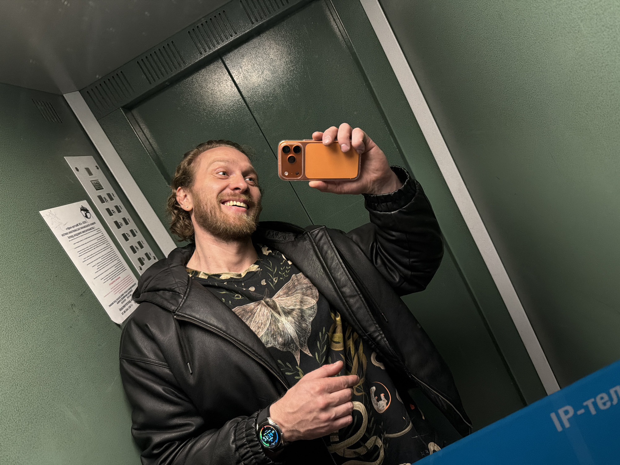 The image shows a smiling man taking a selfie in what appears to be an elevator. He is holding an orange iPhone in his right hand. The man has reddish-blonde hair, a beard, and is wearing a black leather jacket over a patterned shirt. On his left wrist, he is wearing a smartwatch with a visible screen.
The elevator has a green interior with vertical white accents. A notice is affixed to the wall next to a control panel. The notice includes printed text and appears to relate to elevator operations. Part of a blue sign with the Cyrillic text "ІР-тел" is visible in the bottom right corner of the frame.
Based on the Cyrillic text on the notice and sign, the location is likely in Russia. Given the Cyrillic alphabet and general appearance of the interior, it's plausible that the image was taken in Saint Petersburg, Russia.