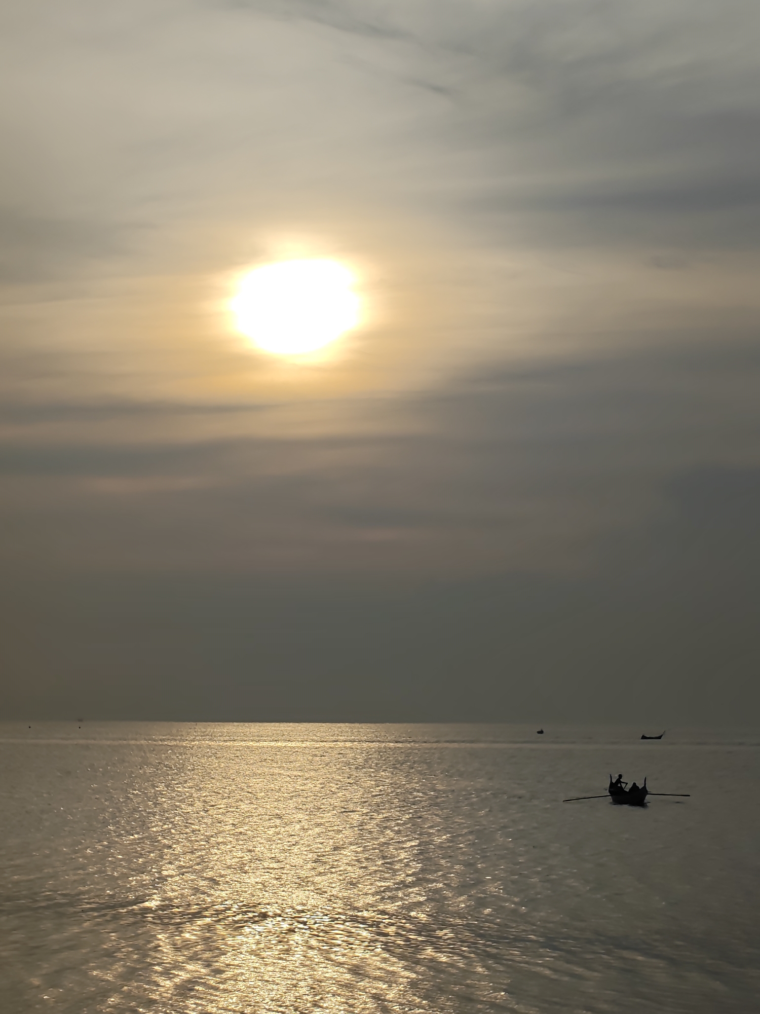 The image presents a serene marine landscape captured in Lhokseumawe, Indonesia, likely during sunrise or sunset. A brilliant, almost blinding, white sun is prominently featured in the upper left quadrant of the frame, casting a golden hue over the entire scene. The sky is partially obscured by soft, hazy clouds, displaying a gradient from light yellow-gray near the sun to darker gray-blue towards the right and top.
The calm water, presumably the sea or ocean, stretches from the foreground to a distant horizon, sparkling intensely with the sun's reflection. The shimmering surface creates a dynamic pattern of golden light and darker ripples.
In the mid-ground, towards the right, a traditional-style boat is seen in silhouette. Two individuals are also silhouetted within the boat, appearing to be rowing, with oars extended outwards. Two other small, dark boat shapes are barely discernible on the distant horizon, suggesting more maritime activity. The overall impression is one of peaceful solitude and the quiet labor of fishing or transport at either dawn or dusk. There is no visible text in the media file.