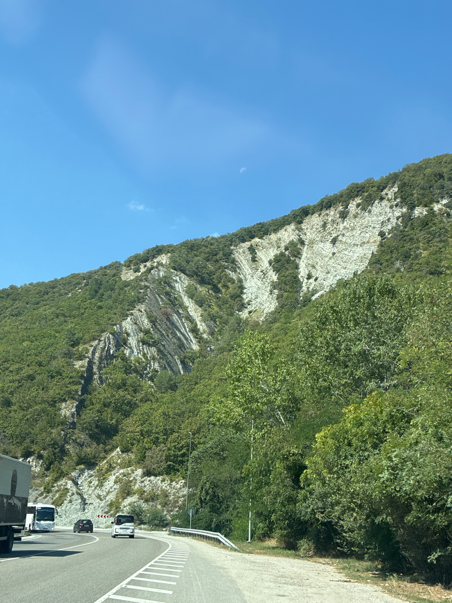 The image captures a scenic view of a road winding through a mountainous area, likely near Shirokaya Schel, Russia. The road is paved and marked with white lines, indicating a highway or major route. Several vehicles are visible on the road: a large truck on the left, followed by a white bus and a black car. Further ahead, a white minivan is seen.
The landscape is dominated by a steep, tree-covered mountain slope on the right side of the road. The mountain features exposed rock formations with distinct layers, suggesting geological activity. The sky is a clear, bright blue with a faint, crescent moon visible. The overall environment suggests a sunny day in a rural, mountainous region.