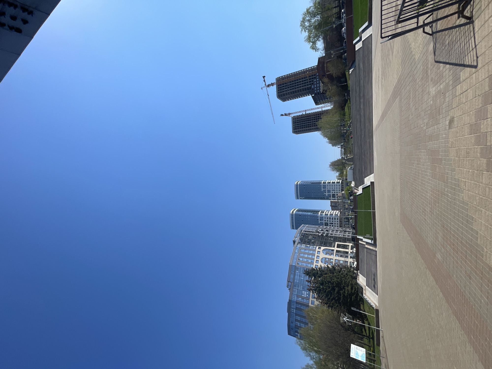 The image depicts a street scene in Izhevsk, Russia, viewed from a low angle. The foreground consists of a paved sidewalk composed of light brown bricks, with a darker brown stripe running parallel to its edge. To the left of the sidewalk are buildings of varying heights and architectural styles, including modern residential towers with glass facades and taller, unfinished buildings under construction, equipped with cranes. Patches of greenery and sparse trees are visible between the buildings. The sky is a clear, bright blue. No people or interactions are clearly visible. The scene appears to be taken on a sunny day.