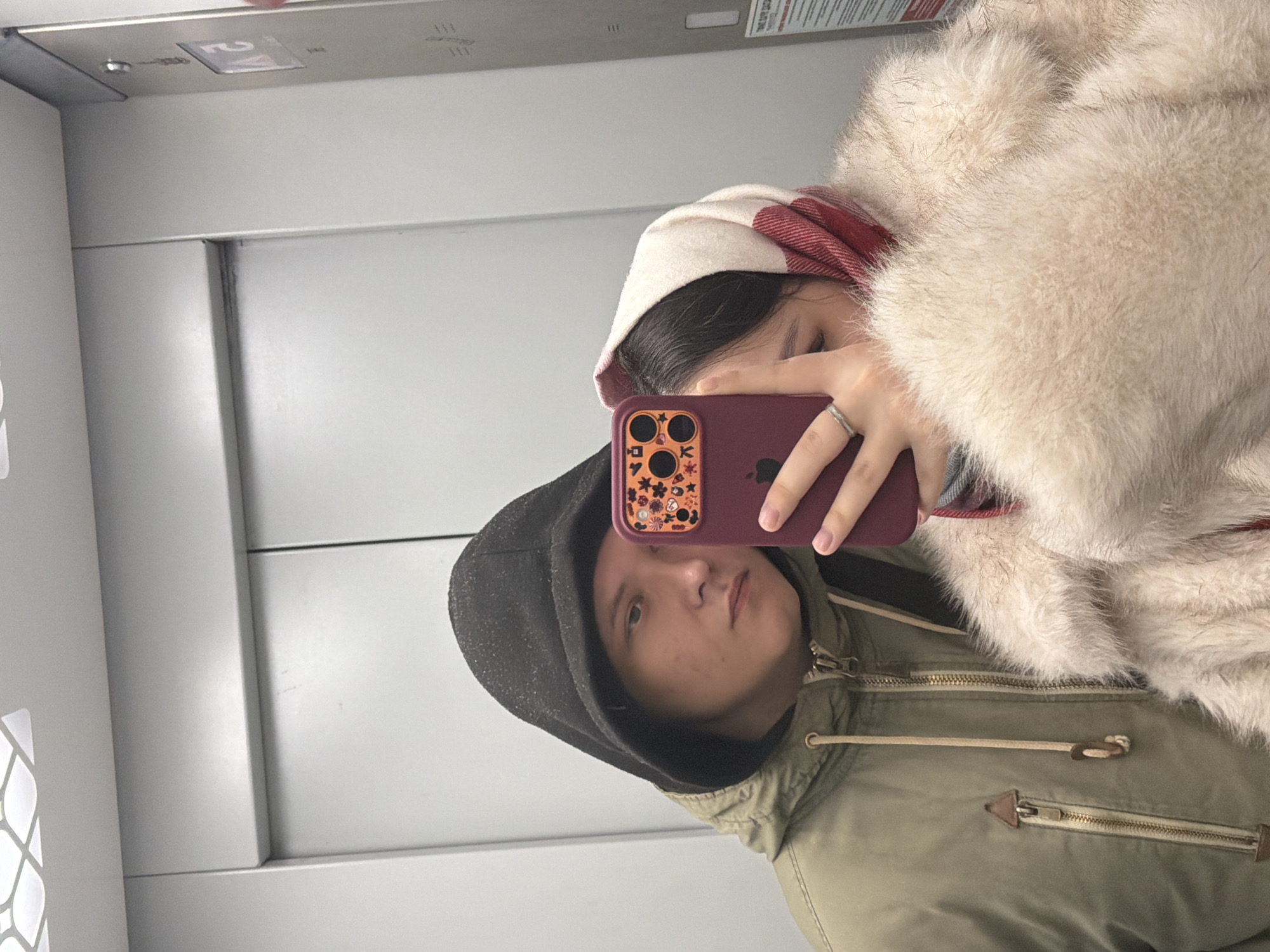 A couple is taking a selfie in what appears to be an elevator. The location is likely in Perm, Russia, as indicated by the elevator panel. The woman is wearing a white and red hat with a large, furry white jacket. She is holding a maroon-colored iPhone with a decorative case. The man is wearing a dark hooded jacket over an olive green jacket. Both are facing the camera, but partially obscured by the phone. The elevator has a simple, grey interior.