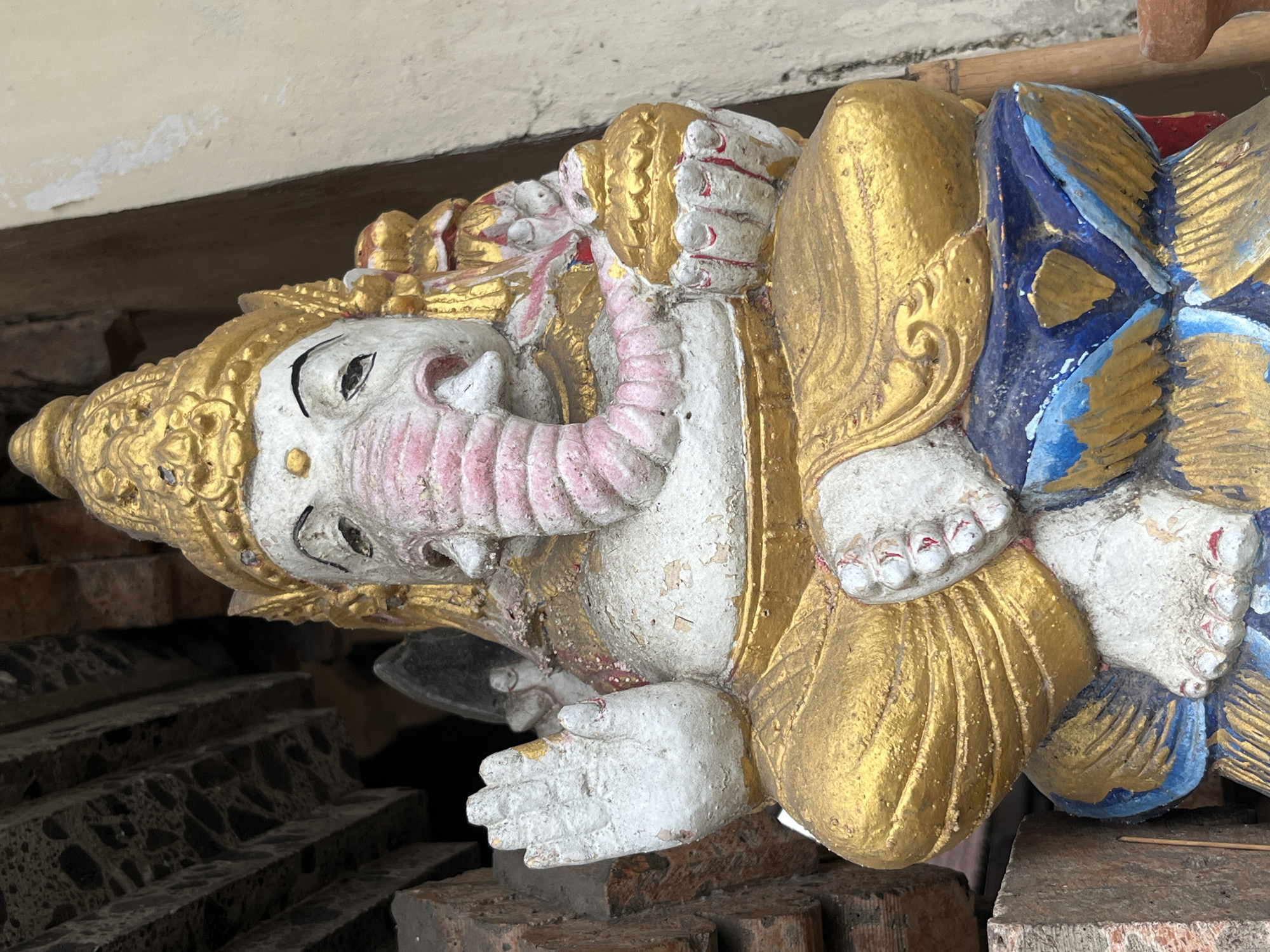 A front-facing, medium shot captures a weathered statue of the Hindu deity Ganesha, positioned outdoors in Kuta Selatan, Indonesia. The statue, seated and facing the camera, is predominantly white and gold, with touches of pink and blue. Ganesha's characteristic elephant head is visible, with a prominent trunk and large ears. The deity's eyes are dark, and a gold, ornate crown adorns its head. In its right hand, Ganesha holds a golden, pot-like object, while its left hand is raised with palm facing outwards, a gesture of blessing or protection. The statue sits on a colorful base, featuring blue and gold embellishments. Behind and to the left of the statue, rustic wooden beams and weathered brickwork are visible, suggesting an architectural context. The lighting appears natural, casting shadows and highlighting the textures of the statue and its surroundings. The overall impression is one of religious significance and aged artistry.