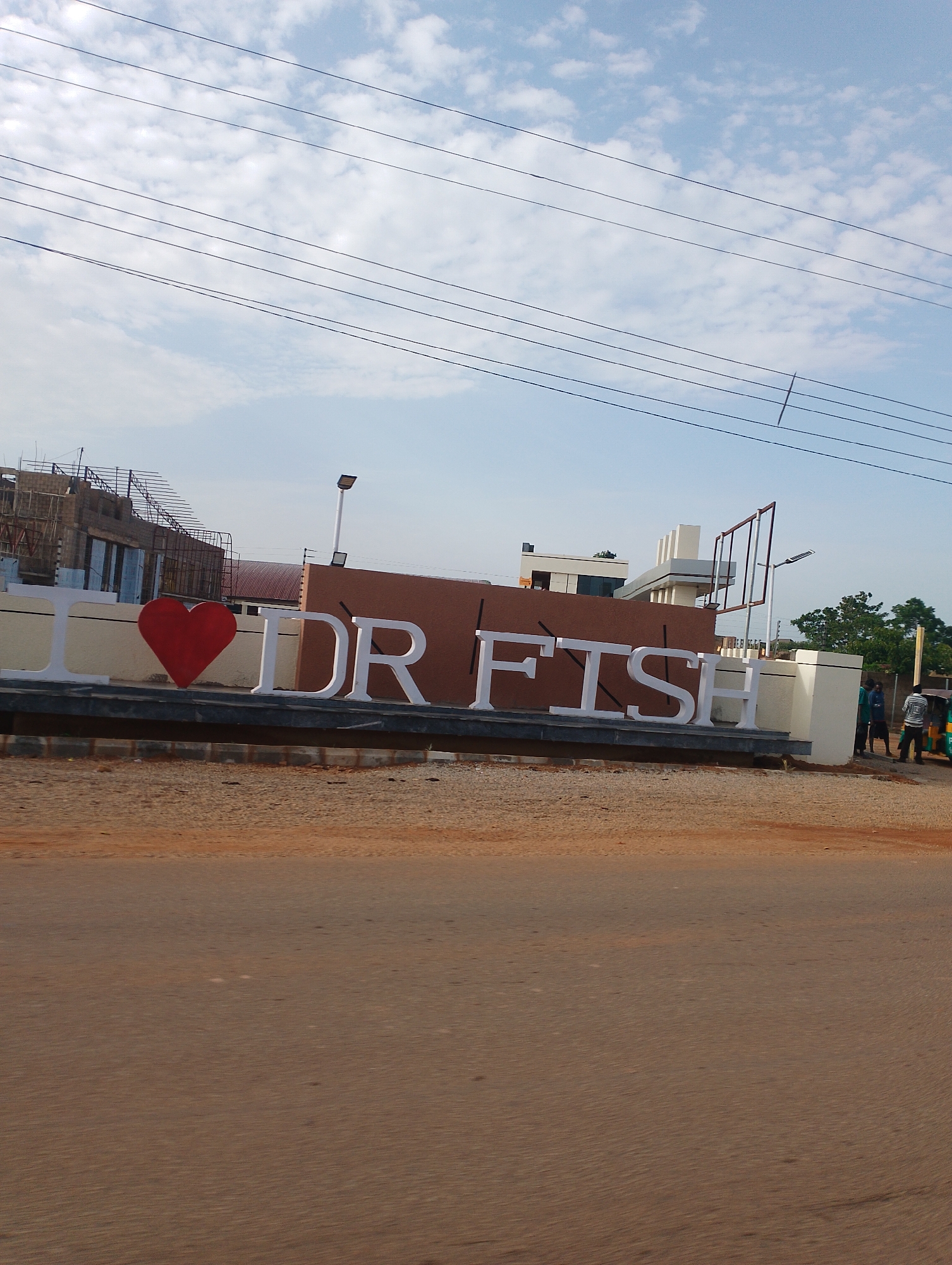 A low-angle, full shot captures a street scene in Gura Top, Nigeria, dominated by a large, white "I LOVE DR FISH" sign. The sign, featuring a red heart, is positioned in the foreground on a dark, raised platform. Behind the sign, construction is underway on a modern building with scaffolding. The background reveals a clear blue sky with scattered clouds and power lines. A few people are visible near the sign and in the distance, along with a three-wheeled auto-rickshaw. The ground in the foreground is composed of dirt and gravel.