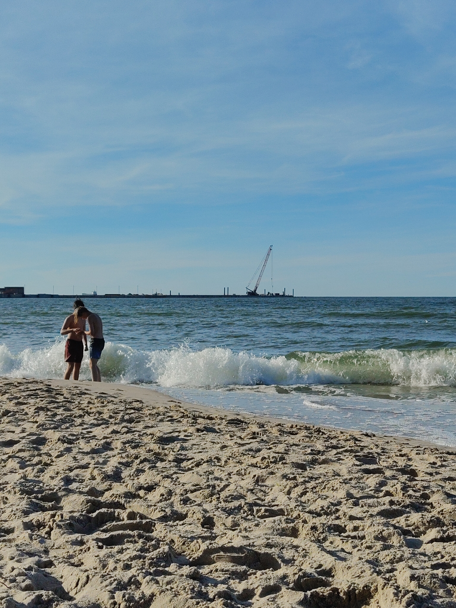 Two individuals are standing on a sandy beach, with waves lapping at their feet. The person on the left is wearing red shorts, and the person on the right is wearing blue shorts. In the background, a large construction crane is visible on the horizon, along with a distant shoreline featuring several structures. The sky is a clear blue with a few wispy clouds. The scene appears to be a coastal area, possibly in Pionersky, Russia, given the presence of the construction crane and industrial-looking structures along the distant coast.