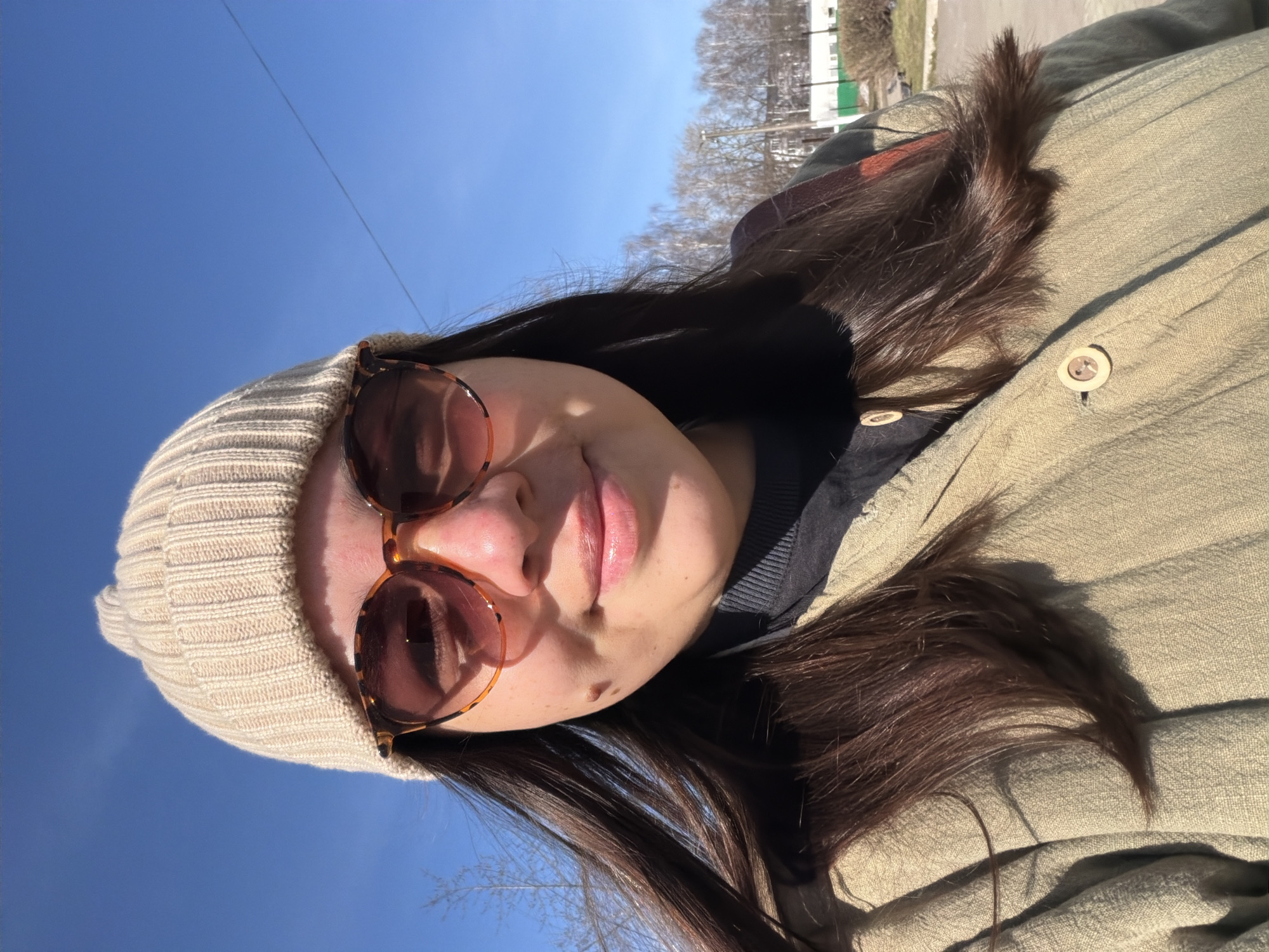 A selfie shows a woman with dark hair and sunglasses. She is wearing a beige beanie and a button-down jacket in a similar color. The sky is blue and visible in the background. Trees and a building are visible in the distant background. The photo was taken in Chaikovsky, Russia.