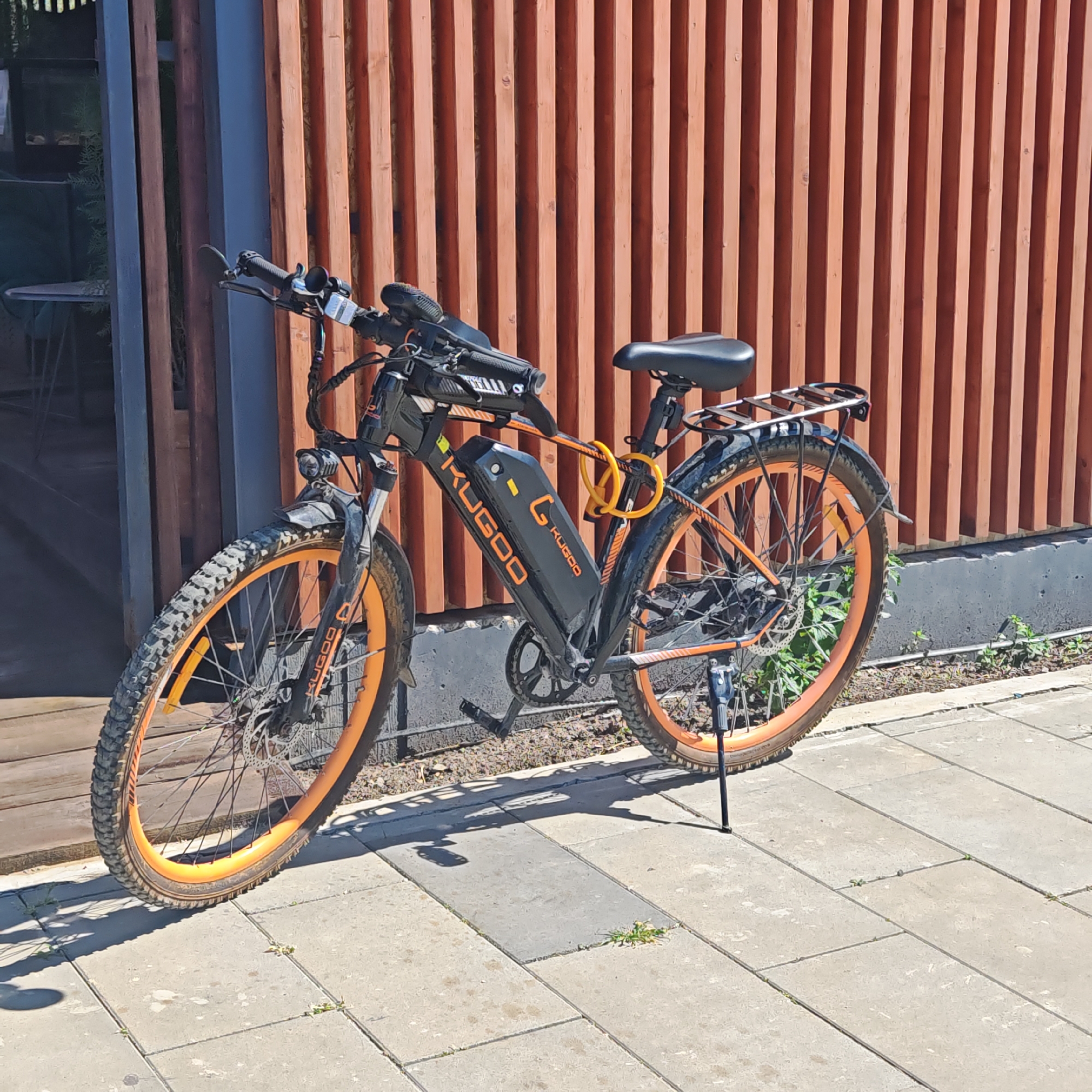 A black Kugoo electric bicycle with orange rims is parked on a light gray paved surface in городское поселение Щелкино, Ukraine. The bicycle leans on its kickstand. It is secured with a yellow U-lock. The background consists of a wooden slatted wall and a partially visible doorway. Some sparse vegetation grows near the base of the wall.