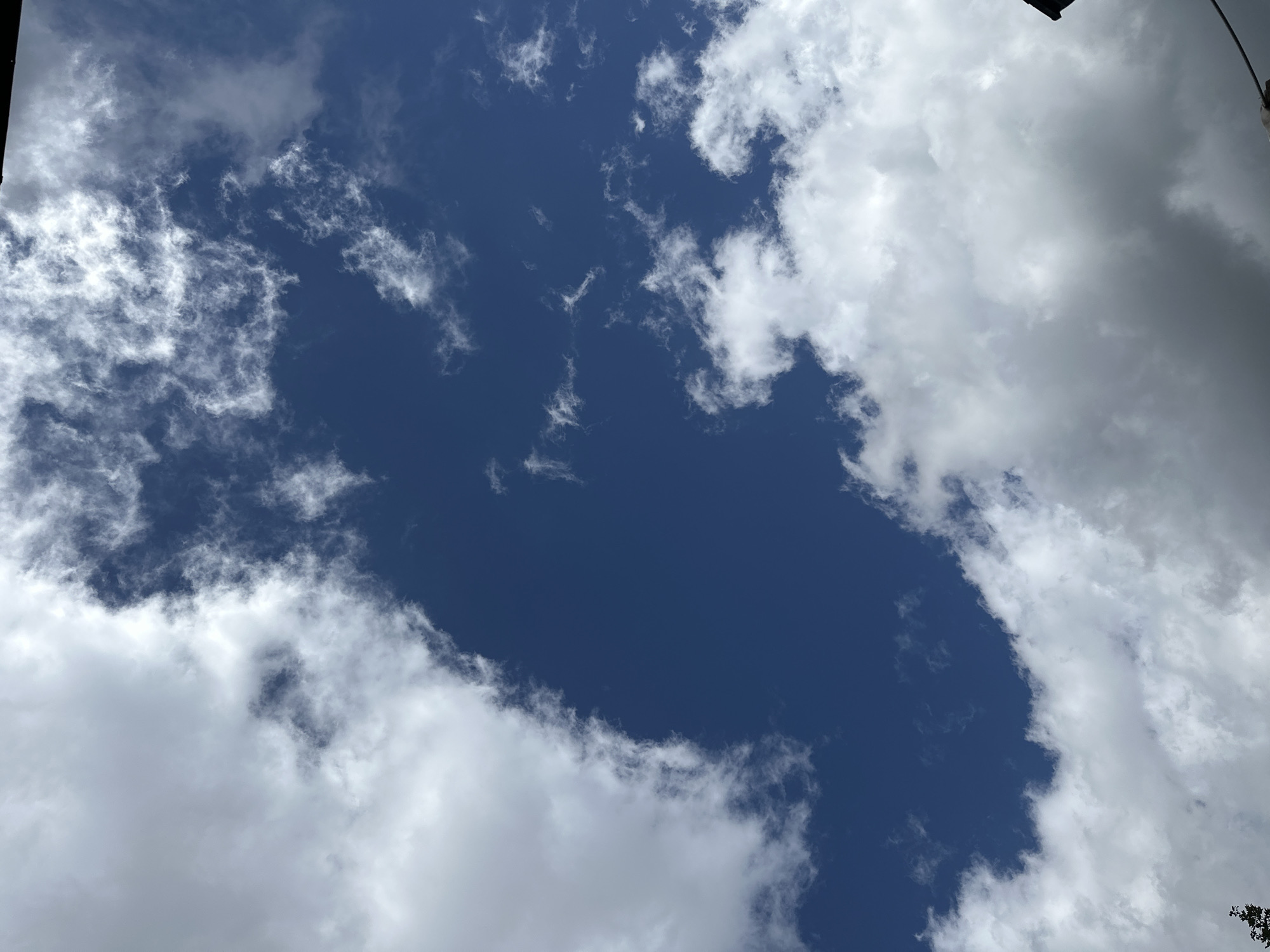 The image shows a deep blue sky partially obscured by white and gray clouds. The clouds are textured, with some appearing wispy and others more dense and puffy. In the lower right corner, a dark silhouette of what appears to be a building or roofline is visible, along with a portion of a thin wire extending horizontally across the frame. The scene depicts an outdoor environment, likely during the daytime given the brightness of the sky and clouds. No people or specific actions are depicted. The location is identified as Corniglio, Italy.