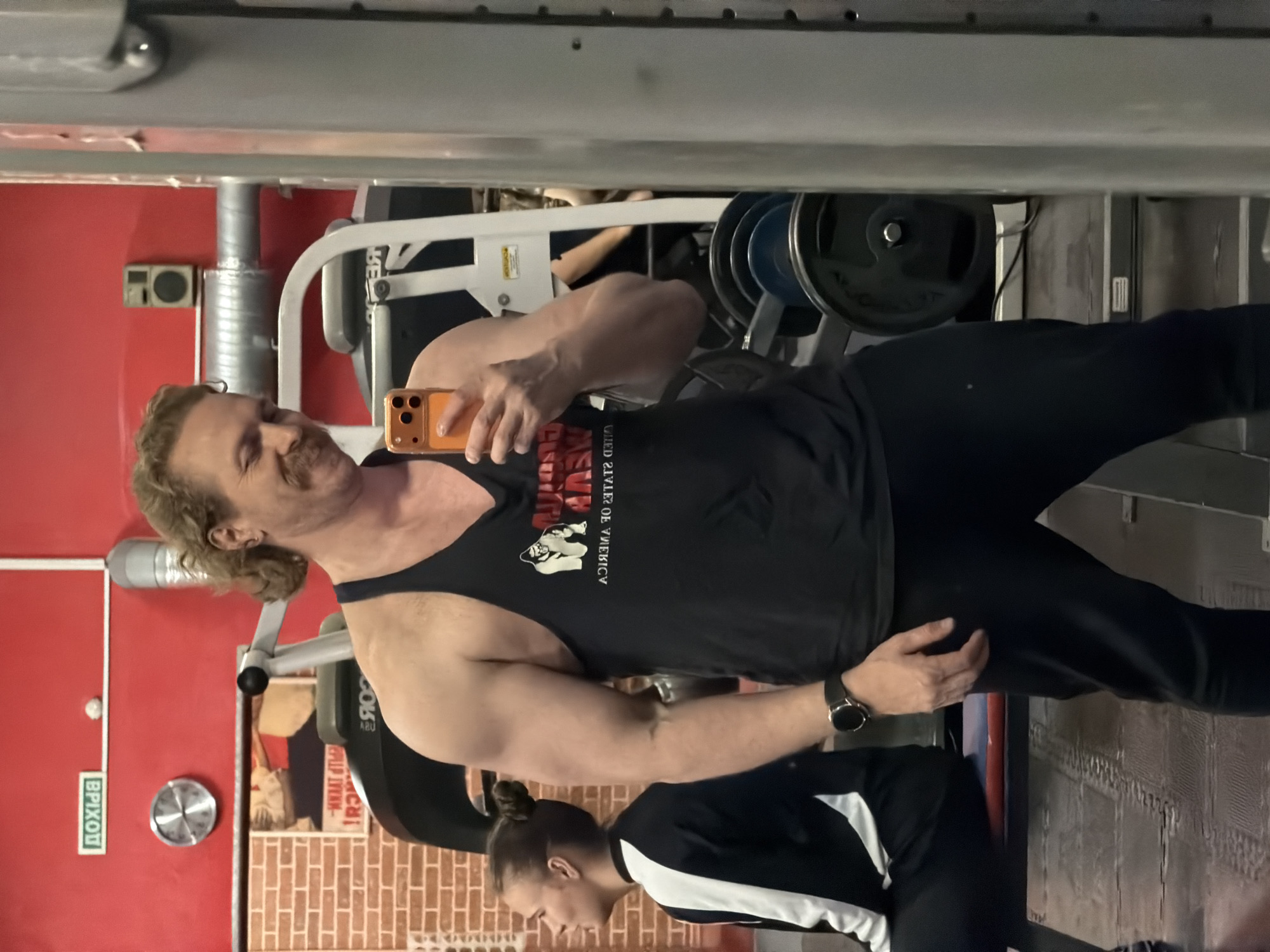 A man is taking a selfie in what appears to be a gym. He's wearing a black tank top and dark pants. He has curly blond hair and a mustache. He is holding a smartphone in his right hand. The gym contains workout equipment, including weight machines. A person with hair in a bun is standing beside him. A sign in Russian is visible on the wall with the word "ВЫХОД" (Exit). The setting appears to be in Saint Petersburg, Russia due to the Russian sign.