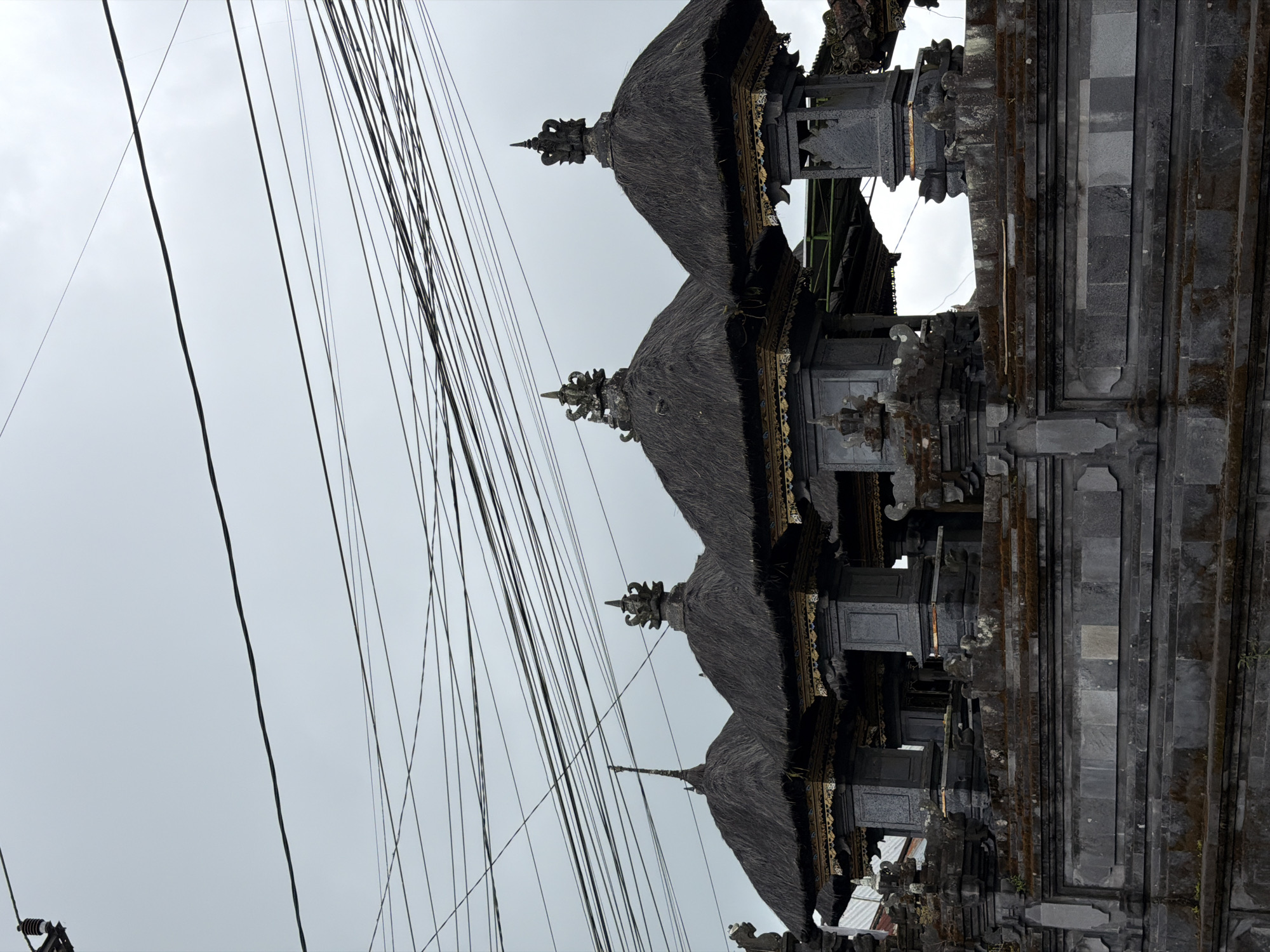 In Kintamani, Indonesia, a series of traditional Balinese structures with thatched roofs and ornate stone pillars are seen. The buildings are aligned along a stone wall, with intricately carved decorative elements on the roofs and pillars. Overhead, a dense network of electrical wires crisscrosses the overcast sky, suggesting an urban or semi-urban setting. The foreground is dominated by the textured stone of the wall and the layered architecture of the buildings, with no people visible in the frame. The overall mood is somber due to the cloudy weather and the imposing, weathered appearance of the structures.