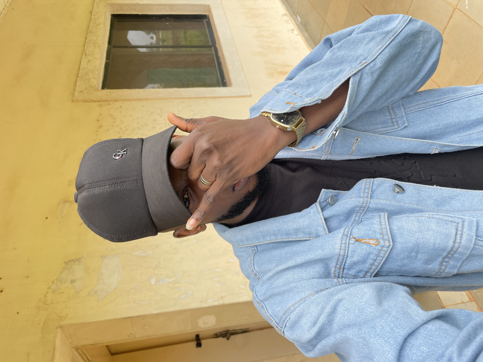 A young Black male is pictured outdoors, wearing a light blue denim jacket over a black t-shirt, a black baseball cap, and a gold wristwatch. He is partially covering his face with his right hand, and he is wearing a ring on his left ring finger. The background appears to be the exterior of a building with yellow-painted walls and a dark-colored window. The scene is likely a residential area in Durumi, Nigeria, given the architectural style and vegetation visible in the background.