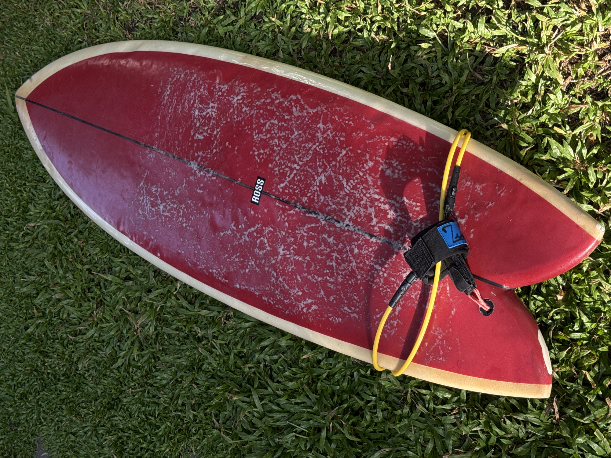 A red surfboard with a light-colored edge and a black stripe down the center lies on green grass. A yellow surf leash is attached to the tail of the board. A black label with white text that reads "ROSS" is visible on the surface of the surfboard. The scene appears to be outdoors in natural light, likely in Goviyapana, Sri Lanka, given the lush green grass and tropical vegetation implied by the lighting and shadows. There are no people or any other objects visible in the image.
