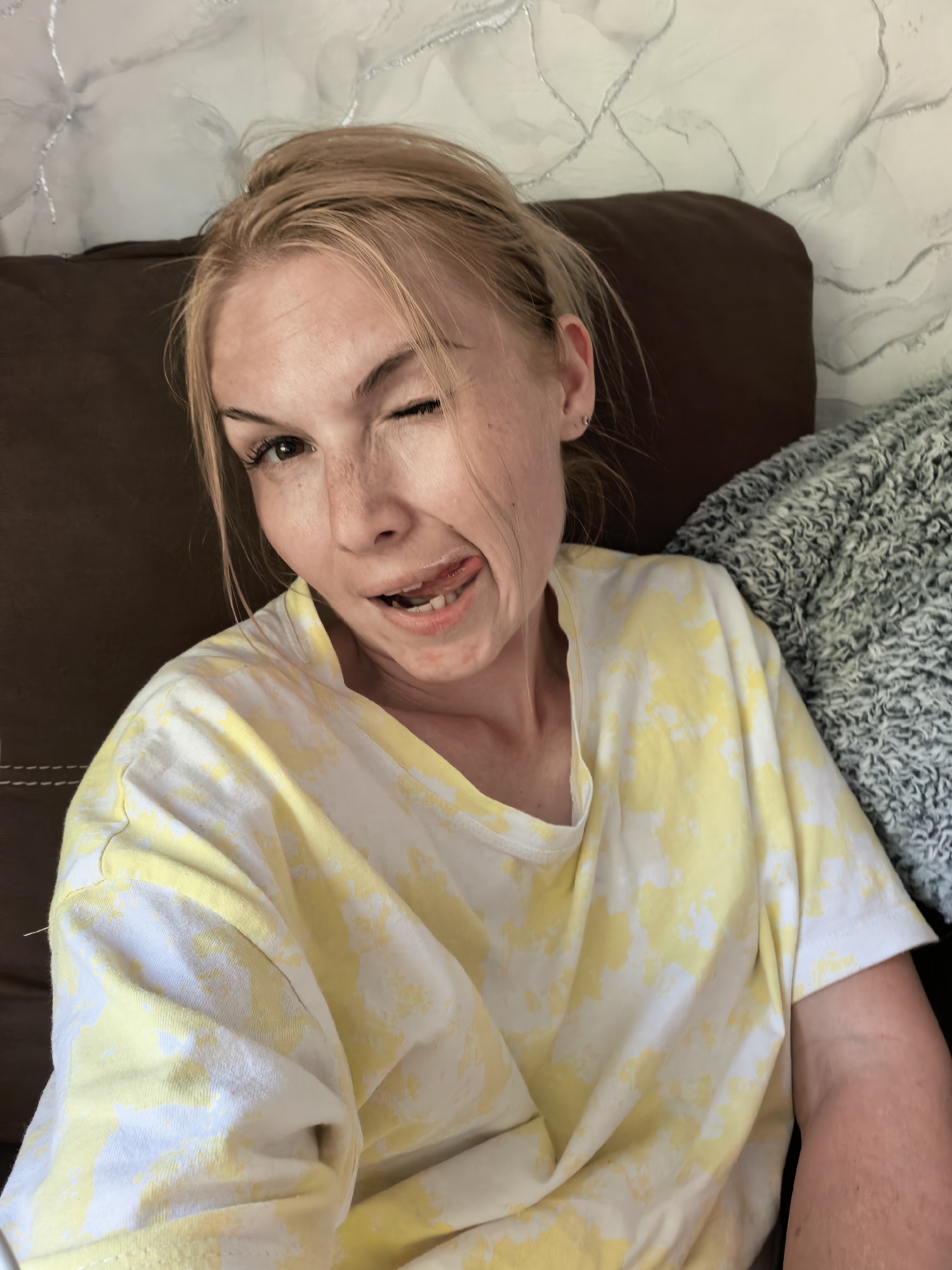 A woman with fair skin and light blonde hair is sitting on a dark brown leather couch. She is wearing a white t-shirt with a yellow tie-dye pattern. She is winking her left eye and sticking her tongue out. Behind her is a white and silver textured wall. A gray knit blanket is partially visible on the right side of the frame. The image does not provide any information about the city of Sterlitamak, Russia.
