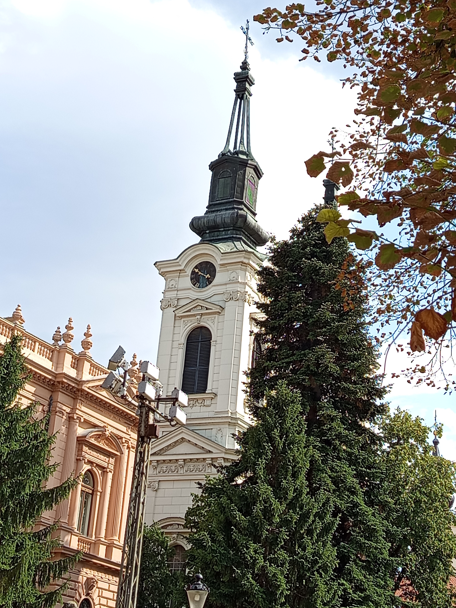 The image captures a view of the Patriarchal Residence in Sremski Karlovci, Serbia. A white church tower with a dark green spire is prominent in the center, featuring a clock face and a tall arched window. To the left of the tower is a section of the pink-hued Patriarchal Residence building, characterized by classical architectural elements such as columns and ornate decorations. In the foreground, a large evergreen tree stands tall, partially obscuring the view of the buildings. Another evergreen tree is visible to the right, and branches with autumnal leaves from a deciduous tree frame the top right portion of the image. The sky above is bright and partly cloudy. In front of the Patriarchal Residence, a metal pole with multiple spotlights is erected.