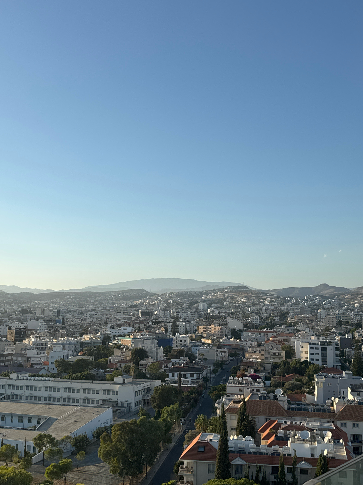 The image shows a panoramic view of the city of Limassol, Cyprus, with a clear blue sky and distant hills. The foreground features a dense urban landscape composed of residential and commercial buildings, characterized by a mix of architectural styles and colors, predominantly white and beige. Several trees are interspersed throughout the city, adding touches of green. A street is visible, with cars parked along its sides and some traffic. The buildings have flat roofs, many adorned with satellite dishes and air conditioning units. In the mid-ground, a large, rectangular building with multiple windows is prominent, likely an institutional or educational facility. The background is dominated by layers of hills and mountains, hazy in the distance, indicating the natural topography surrounding the city. The lighting suggests it is either early morning or late afternoon, with a soft, warm glow illuminating the scene. There are no visible people in the image.