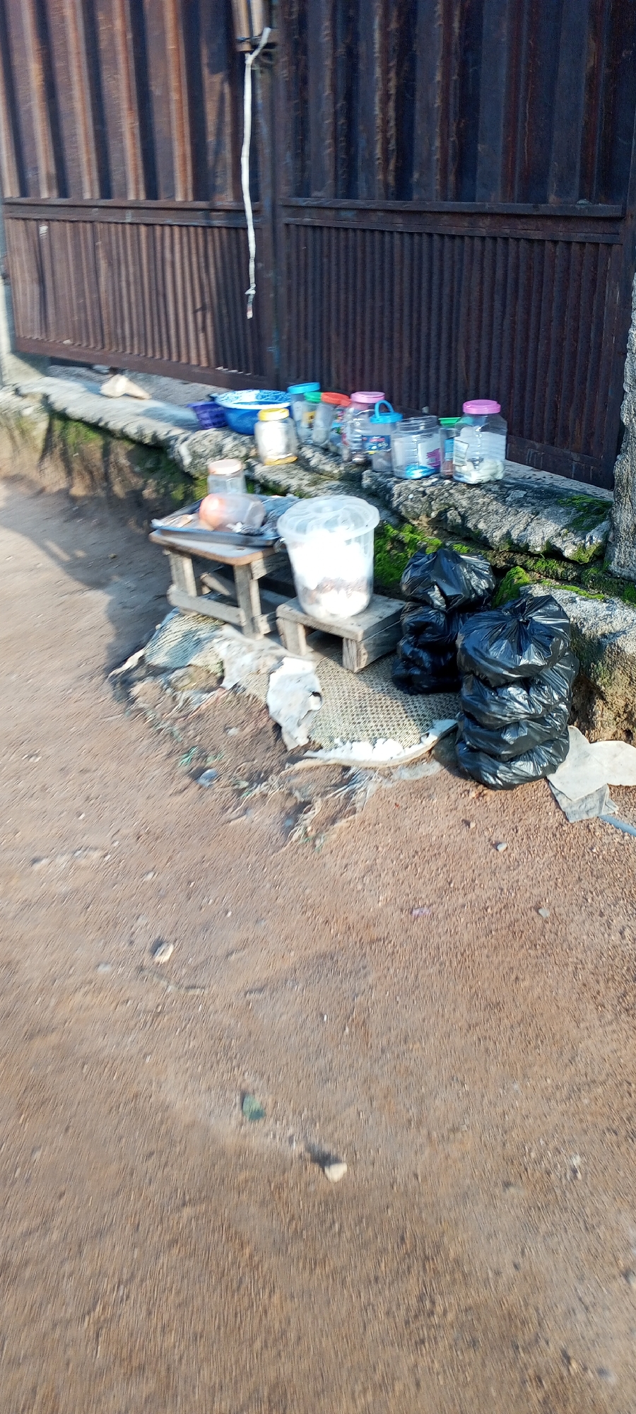 The image depicts an outdoor scene on a dirt path, likely in Jalingo, Nigeria. In the background, a corrugated metal fence, weathered and rusty, forms a backdrop. In front of the fence, a low concrete ledge supports a collection of various items. These include several clear plastic jars and containers of different sizes, some with colorful lids. A blue plastic basin is also visible.
In the foreground, a small wooden stool is placed on the ledge, and on it rests a clear plastic bucket filled with what appears to be a white substance, possibly food. To the right of this, another, smaller wooden stool holds more black plastic trash bags. The ground is a mixture of dirt and gravel, with some scattered debris. The overall impression is of a somewhat unkempt area, possibly near a market or a residential dwelling where items are being stored or sold outdoors.