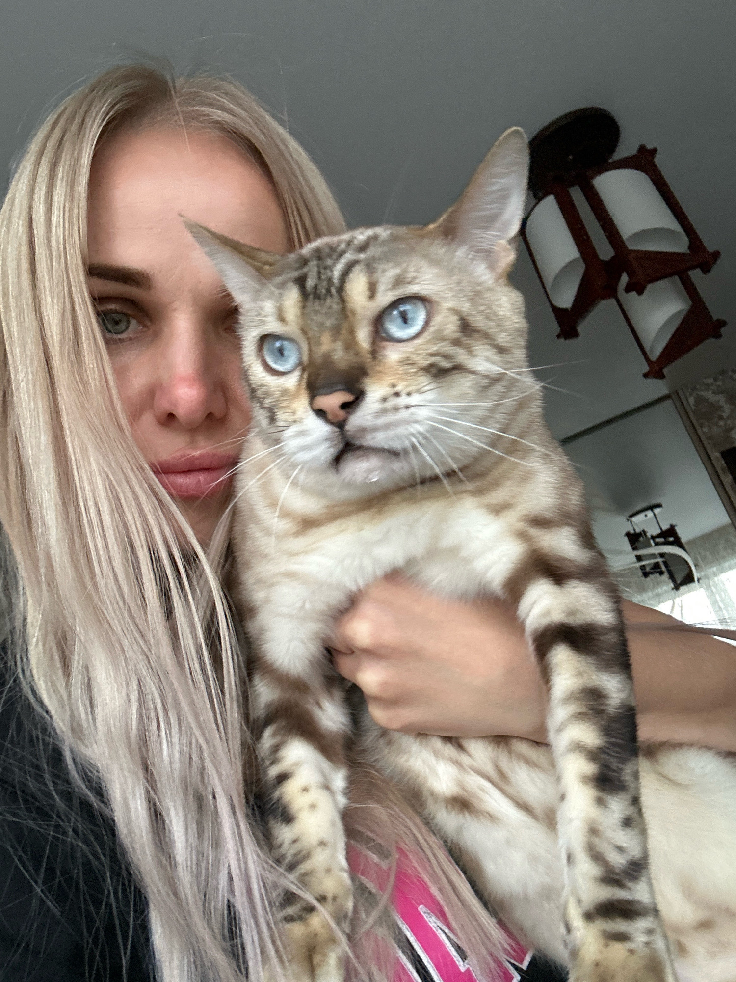 A young woman with blonde hair and striking blue eyes is holding a Bengal cat. The woman is looking towards the camera with a slightly distressed expression, as if she might be crying. The cat, also with blue eyes, is nestled in her arms and looking to its right. The background is out of focus but suggests an indoor setting with a light-colored wall and what appears to be a rustic ceiling lamp. There are no discernible elements that can place the location in Energetik, Russia.