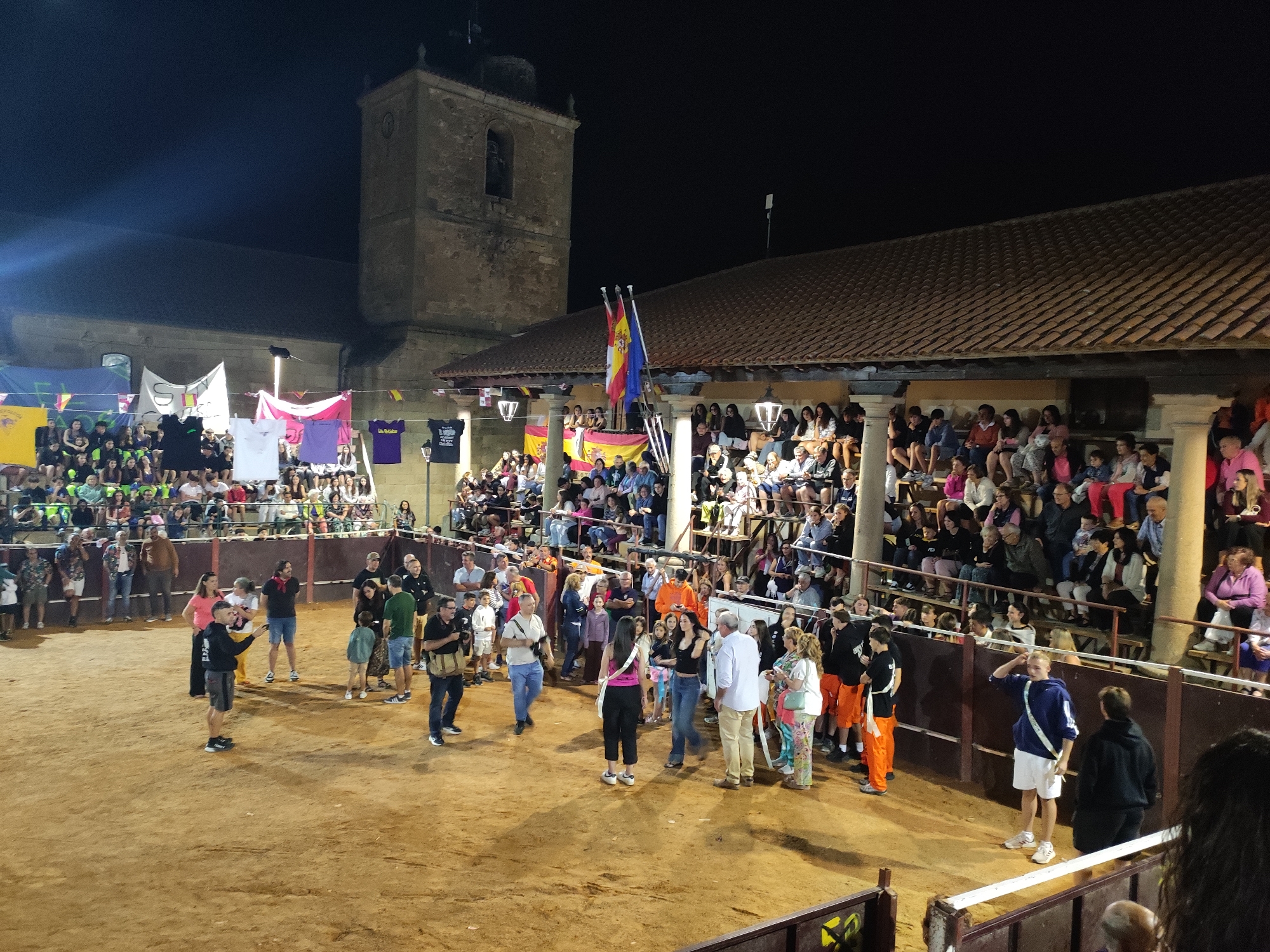 This is a nighttime outdoor photograph taken in the city of Sobradillo, Spain, likely during a festival. The scene depicts a bullring with a dirt floor, surrounded by tiered seating filled with spectators. In the background, a stone tower with a bell is visible, possibly part of a church or other historical building. The event appears to be a public gathering with a festive atmosphere, suggested by the presence of banners and flags, including the Spanish flag. Several people are present in the foreground and middle ground, some standing and observing, while others are seated in the stands. The lighting suggests artificial illumination, likely from floodlights, casting shadows across the arena. The overall impression is one of a community event taking place in a traditional Spanish setting.