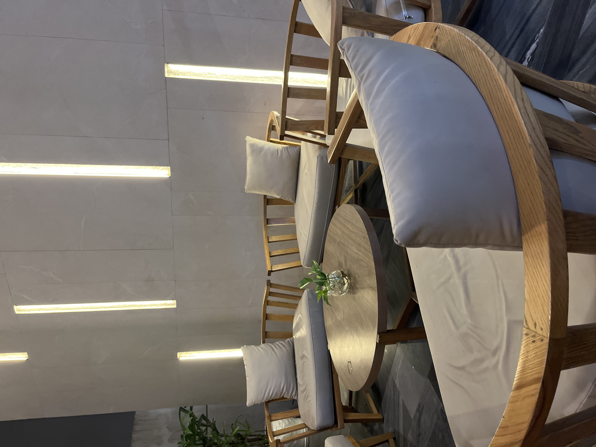 The image shows an indoor seating area with wooden chairs and a round table. The chairs have light-colored cushions and a distinctive curved back. A small vase with greenery sits on the table. The backdrop consists of a wall with linear light fixtures. The floor is patterned in grey. The scene likely depicts a cafe or lounge setting in an urban environment, possibly in Đà Nẵng, Vietnam. No people are visible.