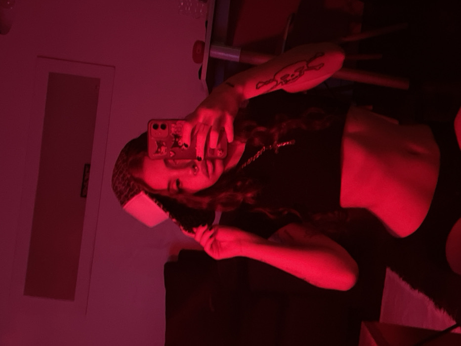 The image displays an indoor scene bathed entirely in red light. A young female individual is positioned centrally, taking a selfie with a smartphone held in her extended right hand. She has long, wavy dark hair and a septum piercing. She wears a dark-colored crop top, revealing her midriff, and dark-colored lower garments that appear to have cut-outs near the hips. Around her neck is a silver-toned chain with a cross pendant. Her left hand holds the brim of a dark-colored baseball cap, worn backward, which features a prominent white panel on the front. A tattoo, depicting a stylized skull and crossed bones or similar emblem, is visible on her right forearm/bicep.
The background shows a light-colored wall, now crimson due to the lighting. An air conditioning unit or similar rectangular fixture is mounted high on the wall. To the left, a dark piece of furniture, possibly a sofa, is partially visible. To the right, a light-colored desk or table is present, holding a clear bottle or glass and another small, indistinct object. The flooring is dark. The specific location within Buenos Aires, Argentina, cannot be determined from the visual evidence presented in the image.