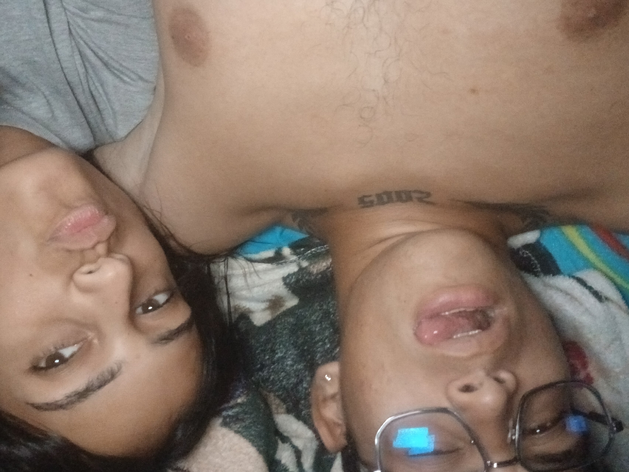 The image shows two individuals lying on a patterned surface, possibly a bed or blanket, with their heads close together. One individual, on the left, has dark hair and is pouting their lips. The other individual, on the right, is wearing glasses and has their mouth slightly open. Part of a bare torso is visible above the head of the individual on the right, with a tattoo that appears to read "2005". The background is a mix of colors and patterns, suggesting a blanket or bedding. There is no information to indicate the location is in Altagracia, Venezuela.