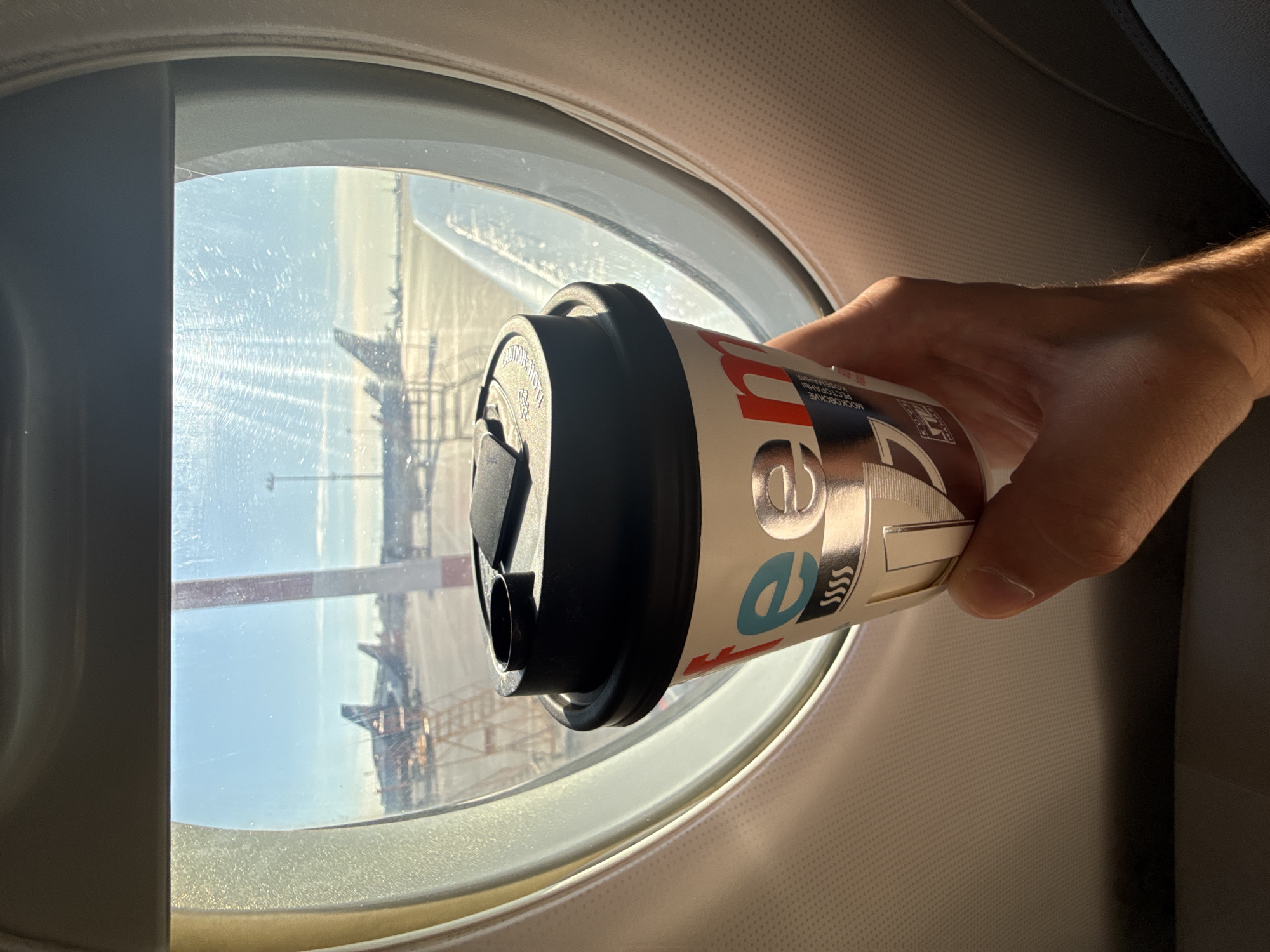 A hand holds a paper coffee cup in front of an airplane window. The cup has a black lid and a white paper sleeve with red, blue, and silver lettering that appears to say "coffee" in a stylized font. The background visible through the window shows an airport tarmac with airplanes, a red and white striped pole, and a bright sky. The interior of the airplane is visible in the foreground, with a textured beige wall. The scene is likely at Sheremetyevo International Airport in Khimki, Russia, given the presence of a coffee cup with Russian text that says "МОСКОВСКИЕ РЕСТОРАНЫ" (Moscow Restaurants) and "КОФЕИ" (Coffee).