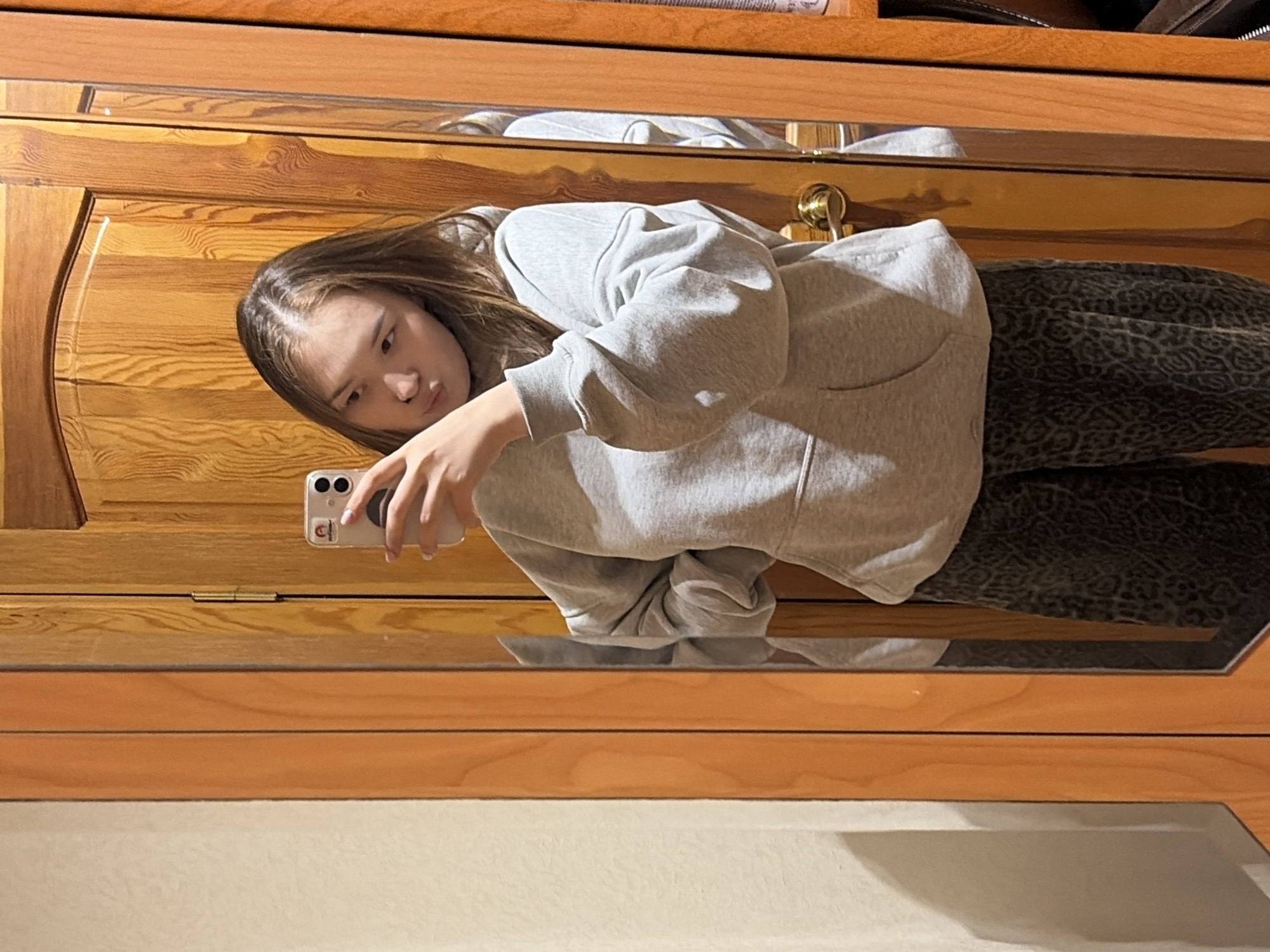 A young woman is taking a selfie in front of a wooden mirrored cabinet or wardrobe. She is wearing a gray hoodie and patterned leggings. The mirror reflects the room, which appears to have light-colored walls. The location is not discernible from the image.