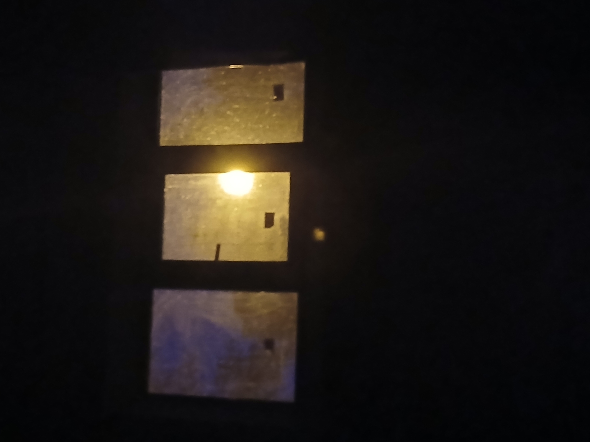 The image displays three rectangular windows set into a dark, solid surface. The windows are slightly reflective, and a bright, yellow light source is visible through the middle window. The light casts a glow that illuminates the surface of the middle and right windows. The overall scene appears to be an exterior view of a dark building at night, possibly a residential building or an industrial facility, located in the city of Ukrainka, Ukraine. There are no people or discernible actions or interactions depicted in the image. The environment is dark, suggesting it is nighttime or the area is not well-lit.