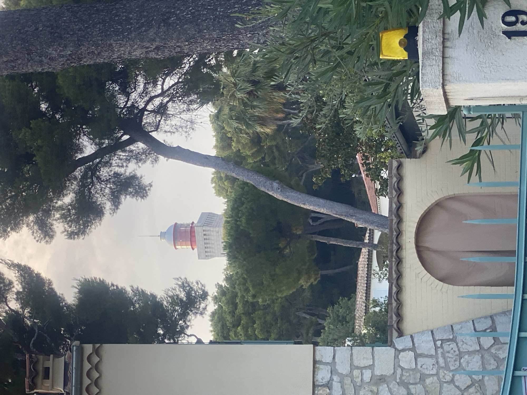 The image depicts a view towards a lighthouse in Antibes, France, partially obscured by dense foliage and trees. In the foreground, a stone wall with a tiled roof section and an arched opening covered by a tan curtain is visible. A teal metal fence with pointed stakes runs across the bottom of the image. To the right, a white pillar supports a yellow flashing alarm light. The number "19" is visible on the pillar. The lighthouse, characterized by its red top and white cylindrical structure, stands in the background, framed by pine trees and other greenery. The sky is overcast.