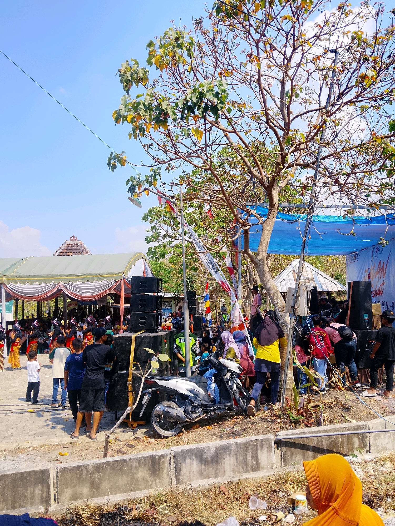 The image depicts an outdoor event taking place in Ngaglik, Indonesia, possibly a cultural festival or a community gathering. In the foreground, a person wearing a bright orange hijab is partially visible, facing away from the camera. Behind them, dry grass and a concrete barrier line the edge of the scene.
The midground is bustling with activity. A large tree with sparse foliage dominates the right side, with wires and flags strung between its branches. At the base of the tree, a stack of large black speakers is prominent, with a white motorcycle parked nearby. A group of people are gathered around the speakers and the motorcycle. To the left of the tree and speakers, a long white tent with decorative drapes is set up, providing shade for a seated audience. Children and adults in casual attire are visible under the tent. Some children are standing in front of the tent, facing towards the main event area. In the background, a building with a tiered, tiled roof, characteristic of Indonesian traditional architecture, is visible. The sky is a clear blue with a few wispy clouds. The overall impression is one of a lively, public gathering under a sunny sky.