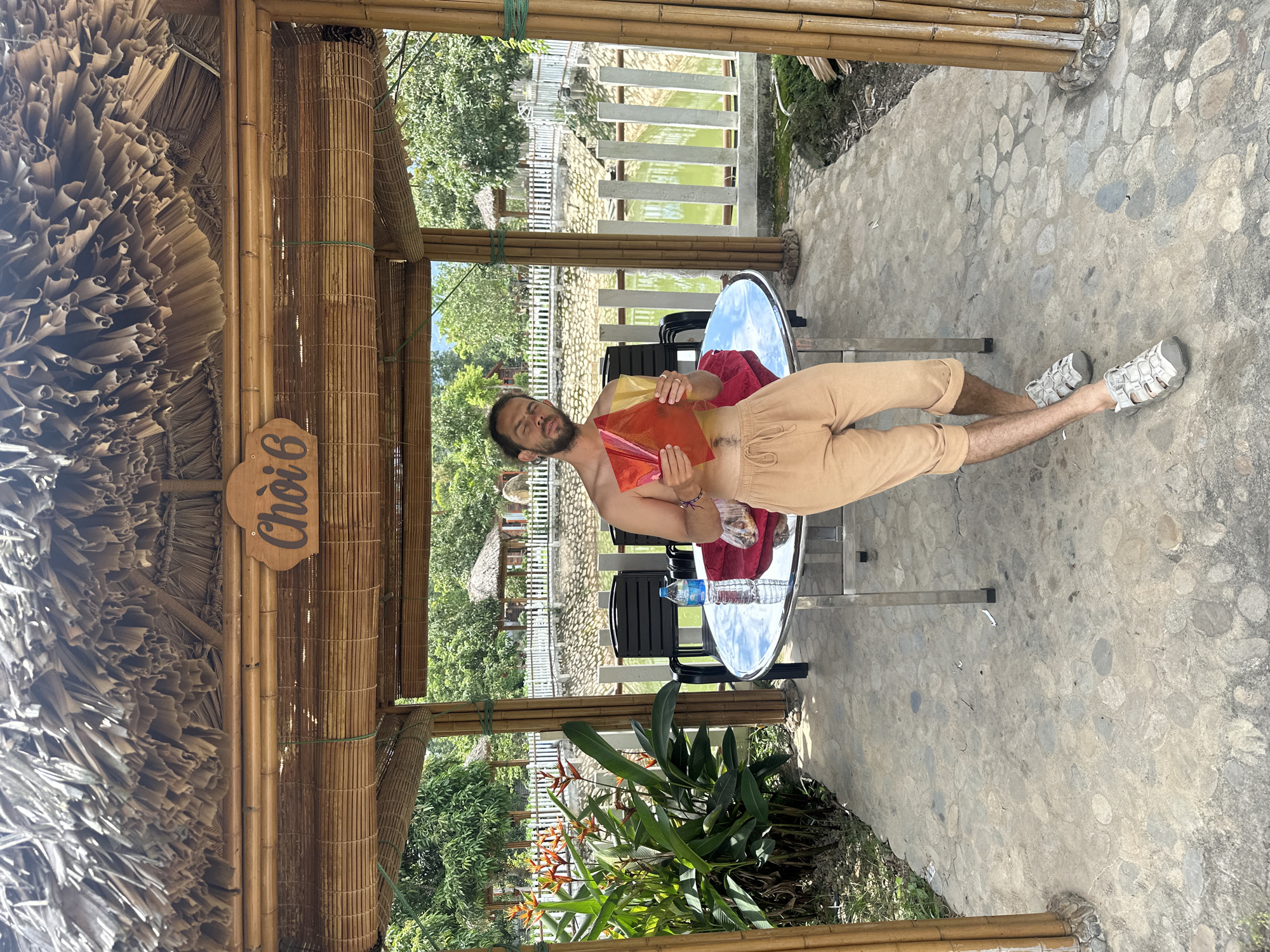 The scene depicts an outdoor setting in Nghia Lo Ward, Vietnam, under a bamboo-framed structure with a thatched roof. A wooden sign reading "Chòi 6" is affixed to the structure's frame. A lone adult male, with a beard and mustache, stands shirtless and wears light brown drawstring pants that extend to mid-calf, paired with light grey closed-toe sandals. He leans against a round table with a reflective surface and a metal base. In his hands, he holds two translucent rectangular sheets, one red and one yellow. On the table, a plastic bottle of water with a blue cap and a crumpled red fabric item are visible. Behind the table, two black chairs are present. The ground beneath the structure is paved with irregular grey stones set in concrete. To the left, a cluster of green plants with orange Heliconia flowers grows. In the background, a rectangular pond or canal with green water is bordered by a white picket fence. Beyond the pond, additional thatched-roof structures and dense green foliage are visible under a clear sky, indicating a sunny daytime environment.