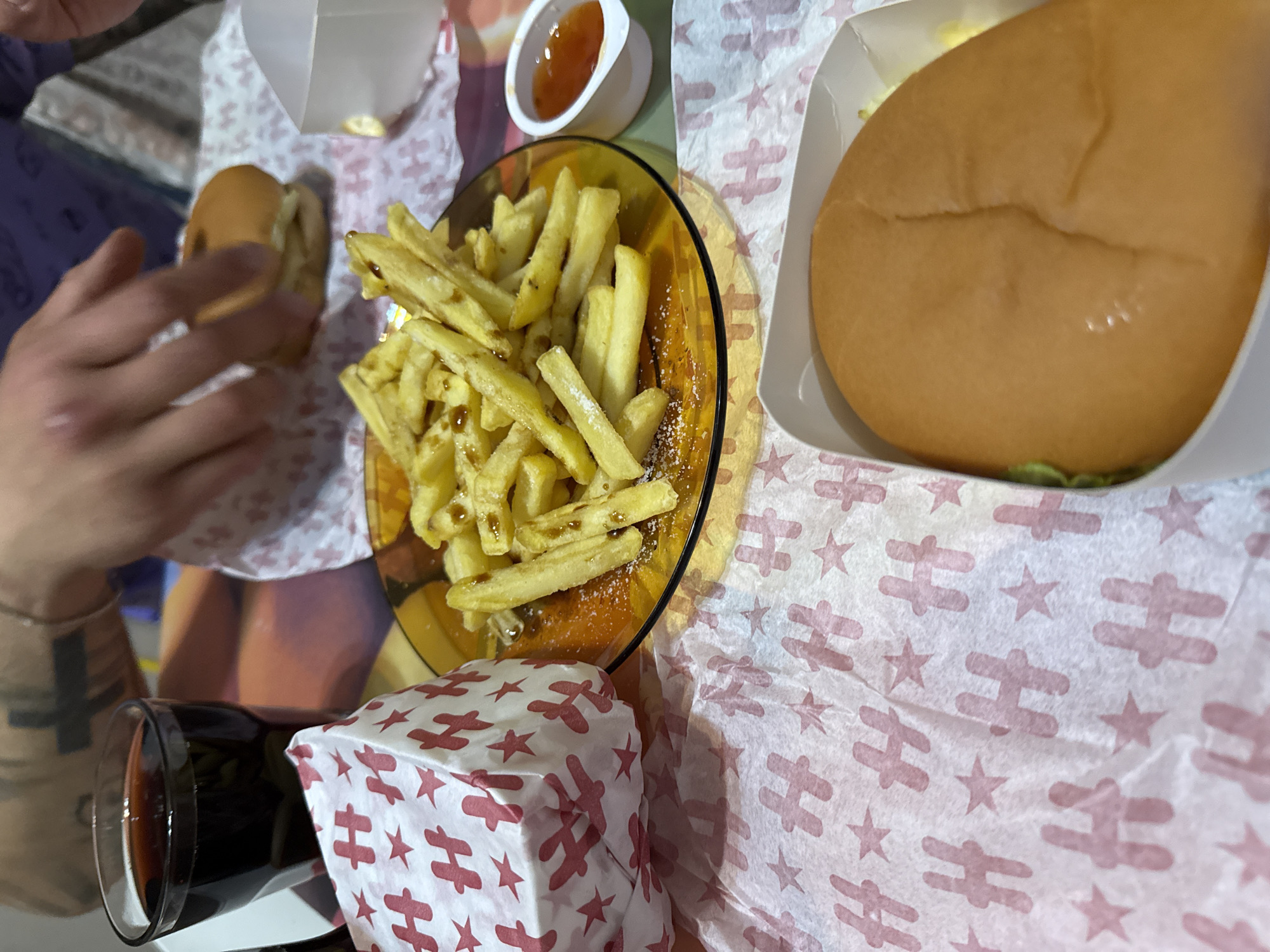 The image shows a table setting with fast food items. There is a burger in a white paper container, a glass plate of French fries with some sauce on them, a small container of dipping sauce, a paper-wrapped item with a red and white airplane and star pattern, a glass of dark liquid (possibly cola), and a person's hands holding a small sandwich. The food and drink are placed on a white paper with a red pattern of stars and airplanes. The specific location in Varna, Bulgaria cannot be determined from this image.